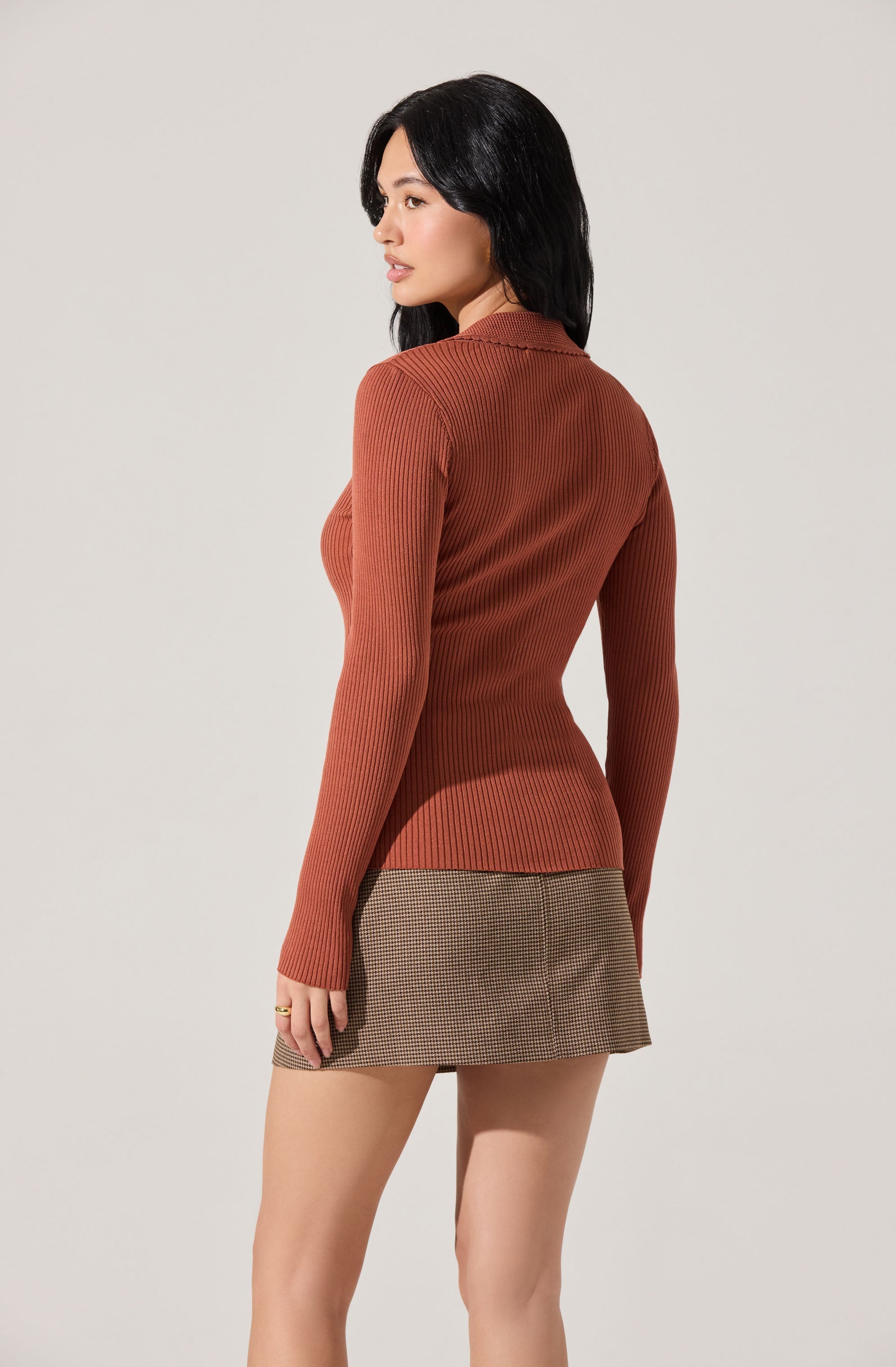 A woman is wearing a fitted, long-sleeved, ribbed sweater in a rust color. The sweater has a collar and is tucked into a houndstooth patterned mini skirt in brown and beige tones.