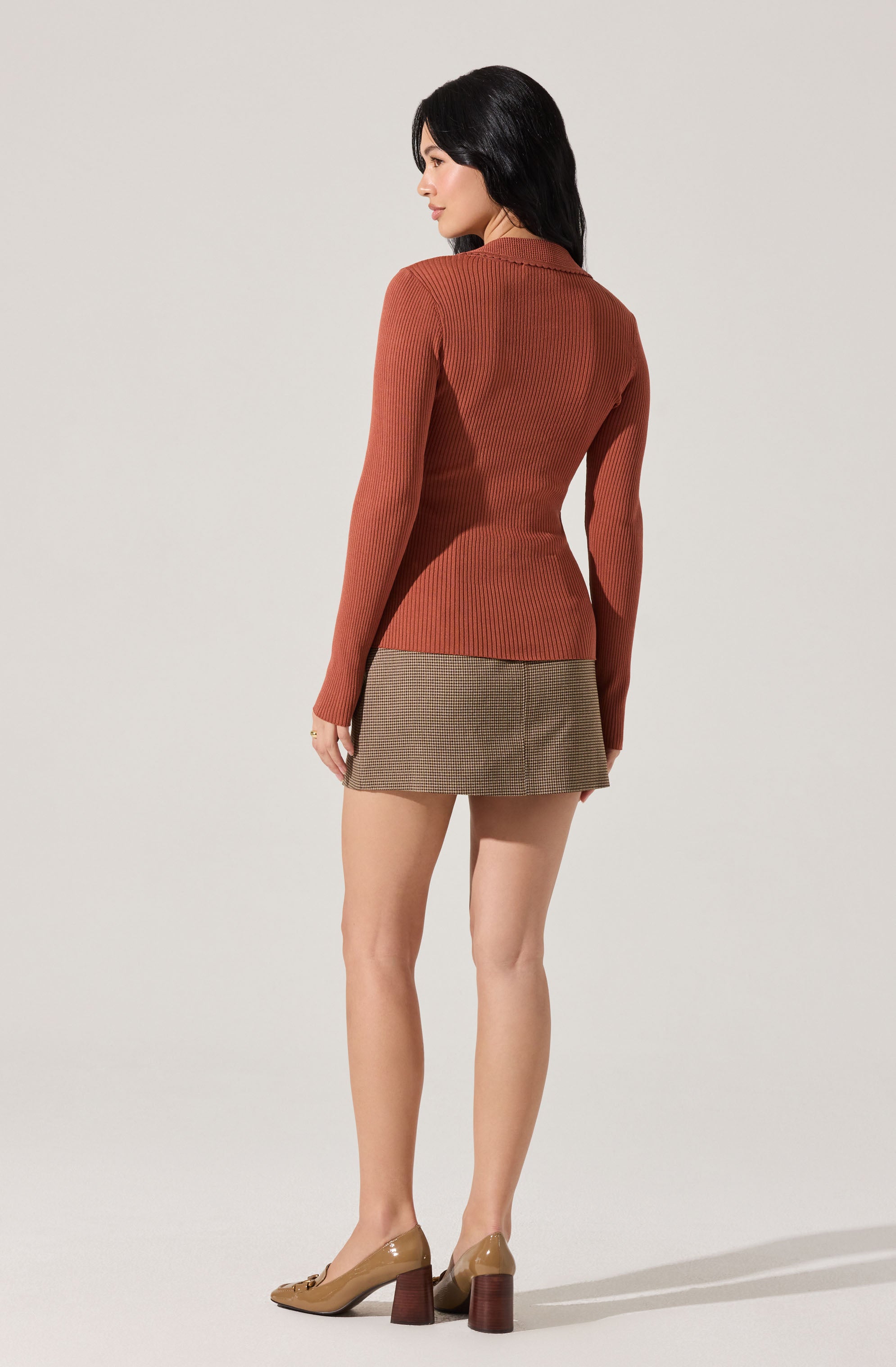 A woman is wearing a fitted, long-sleeved, ribbed sweater in a rust color. She pairs it with a short, patterned skirt in shades of brown and beige. She completes her outfit with tan high-heeled shoes that have a chunky, dark brown block heel.