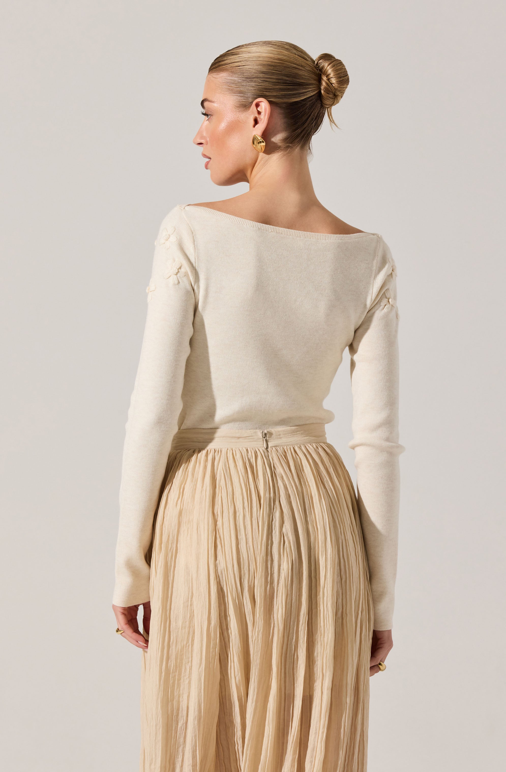 A woman is wearing a long-sleeved, off-white sweater with subtle textured detailing on the shoulders. She pairs this with a high-waisted, beige, pleated skirt that has a slight sheen and a zipper closure at the back. She is accessorized with a pair of gold earrings and rings.