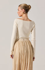 A woman is wearing a long-sleeved, off-white sweater with subtle textured detailing on the shoulders. She pairs this with a high-waisted, beige, pleated skirt that has a slight sheen and a zipper closure at the back. She is accessorized with a pair of gold earrings and rings.