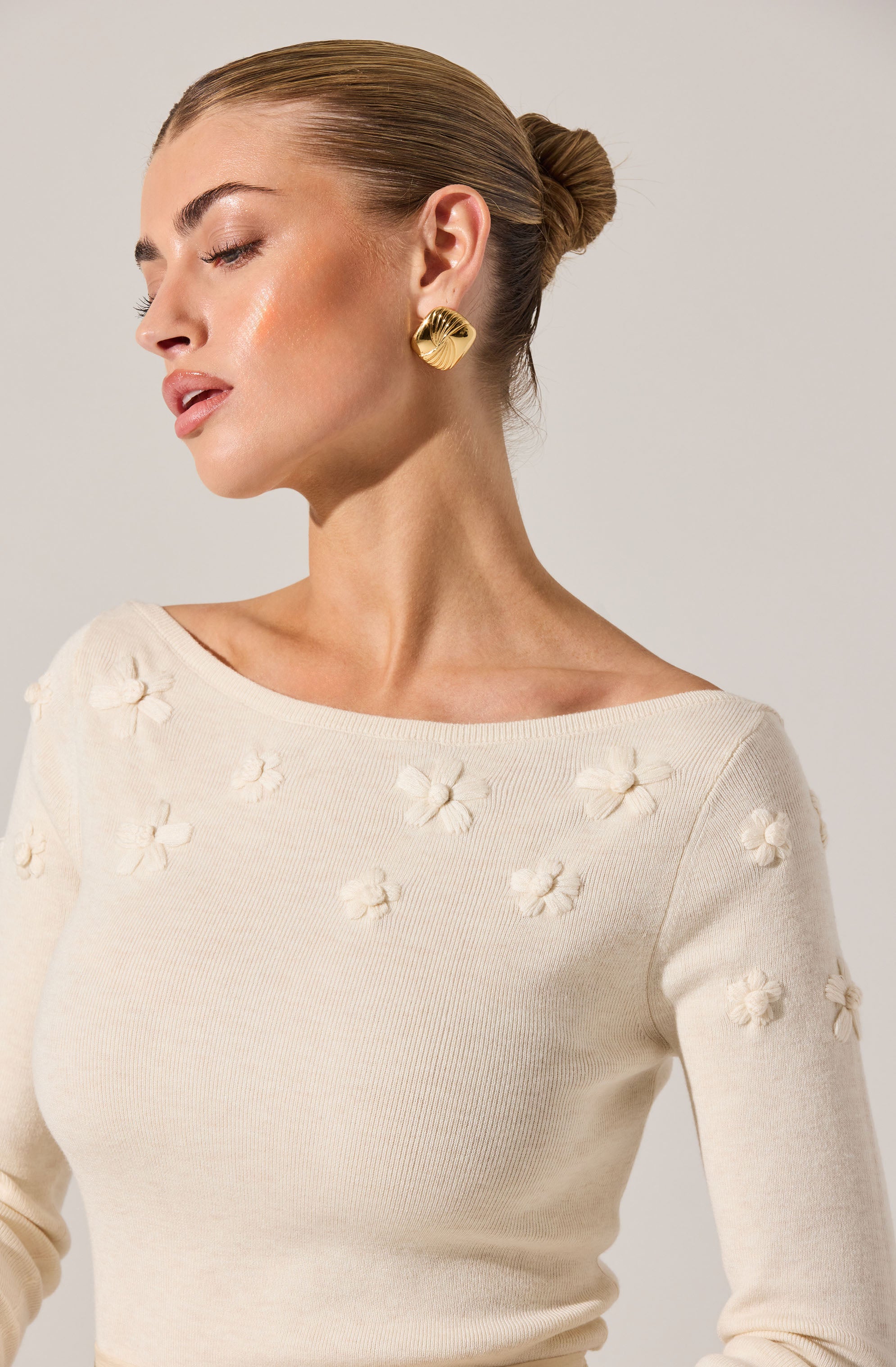 A woman is wearing a cream-colored sweater with a boat neckline. The sweater features small, raised floral patterns scattered across the fabric.