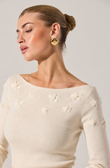 A woman is wearing a cream-colored sweater with a boat neckline. The sweater features small, raised floral patterns scattered across the fabric.