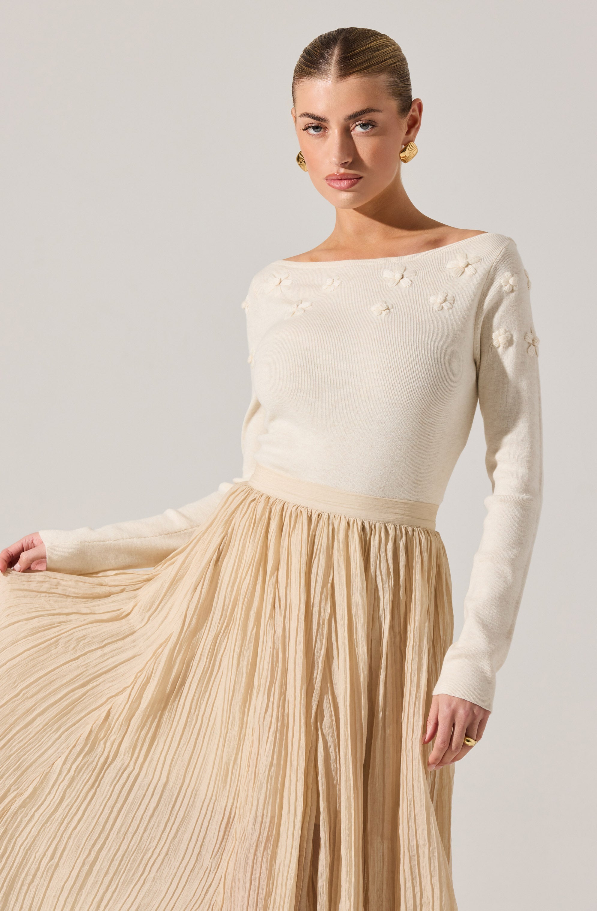 A woman is wearing a long-sleeved, cream-colored sweater with small floral embellishments on the shoulders and upper chest. The sweater is tucked into a high-waisted, beige pleated skirt that reaches down to her ankles.