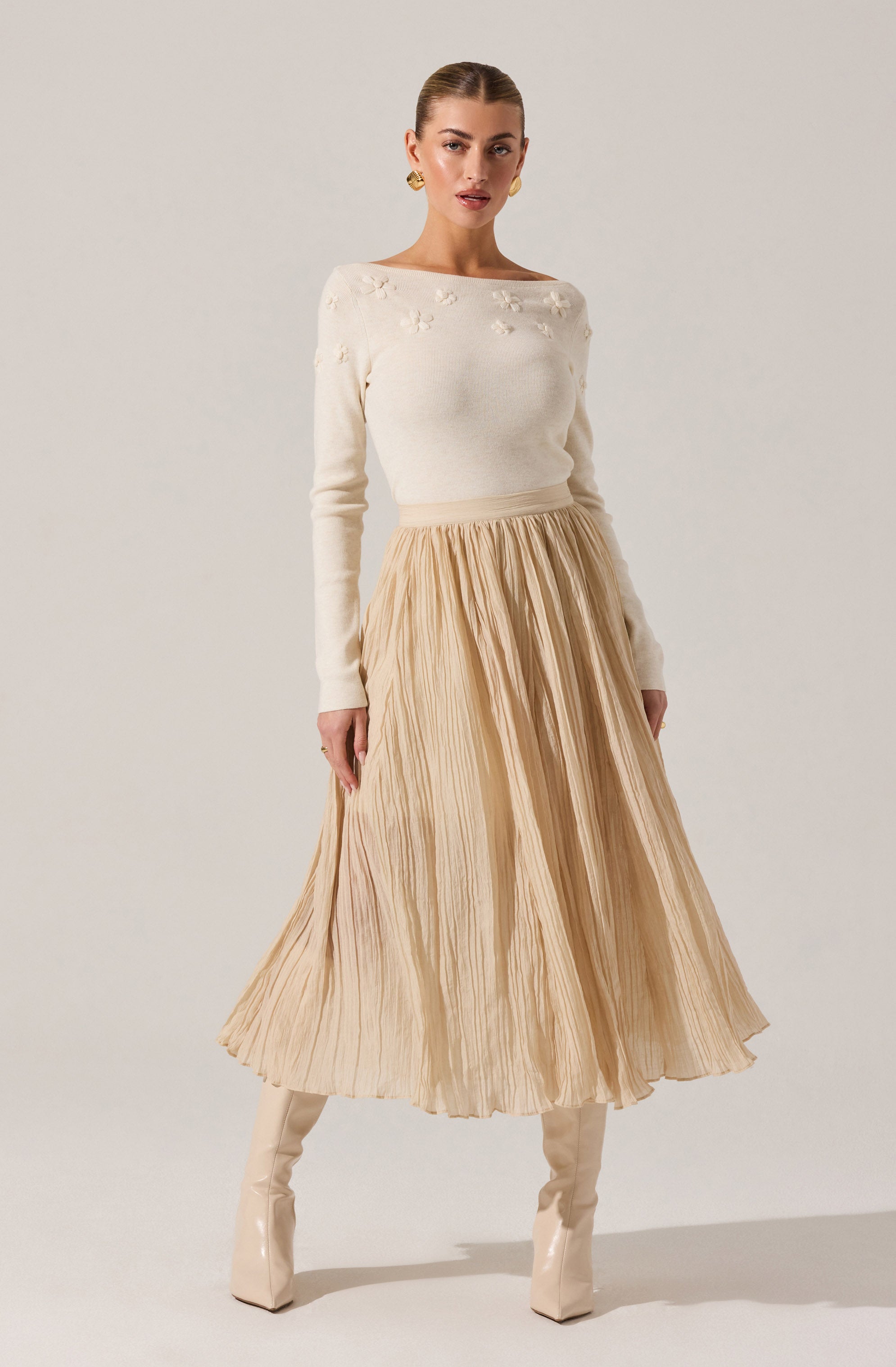 A woman is wearing an off-white, long-sleeved sweater with textured floral embellishments on the chest area. She pairs it with a high-waisted, pleated midi skirt in a light beige or champagne color, and matching beige knee-high boots. She has on gold hoop earrings and her hair is styled in a sleek, pulled-back look.