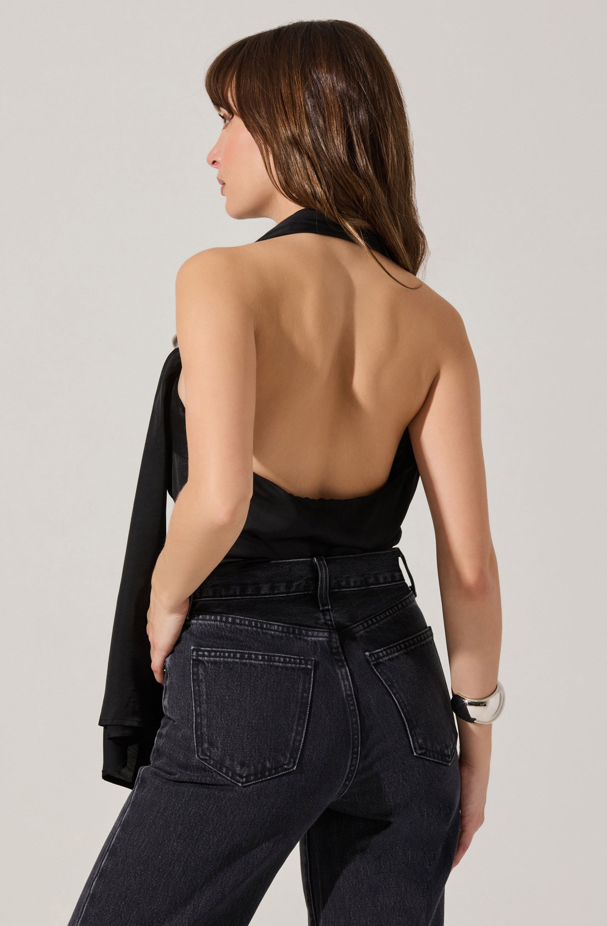 A woman is wearing a sleeveless, backless black top and high-waisted black jeans. She has a silver wristwatch on her left wrist and is holding a black jacket over her left arm.