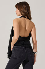 A woman is wearing a sleeveless, backless black top and high-waisted black jeans. She has a silver wristwatch on her left wrist and is holding a black jacket over her left arm.