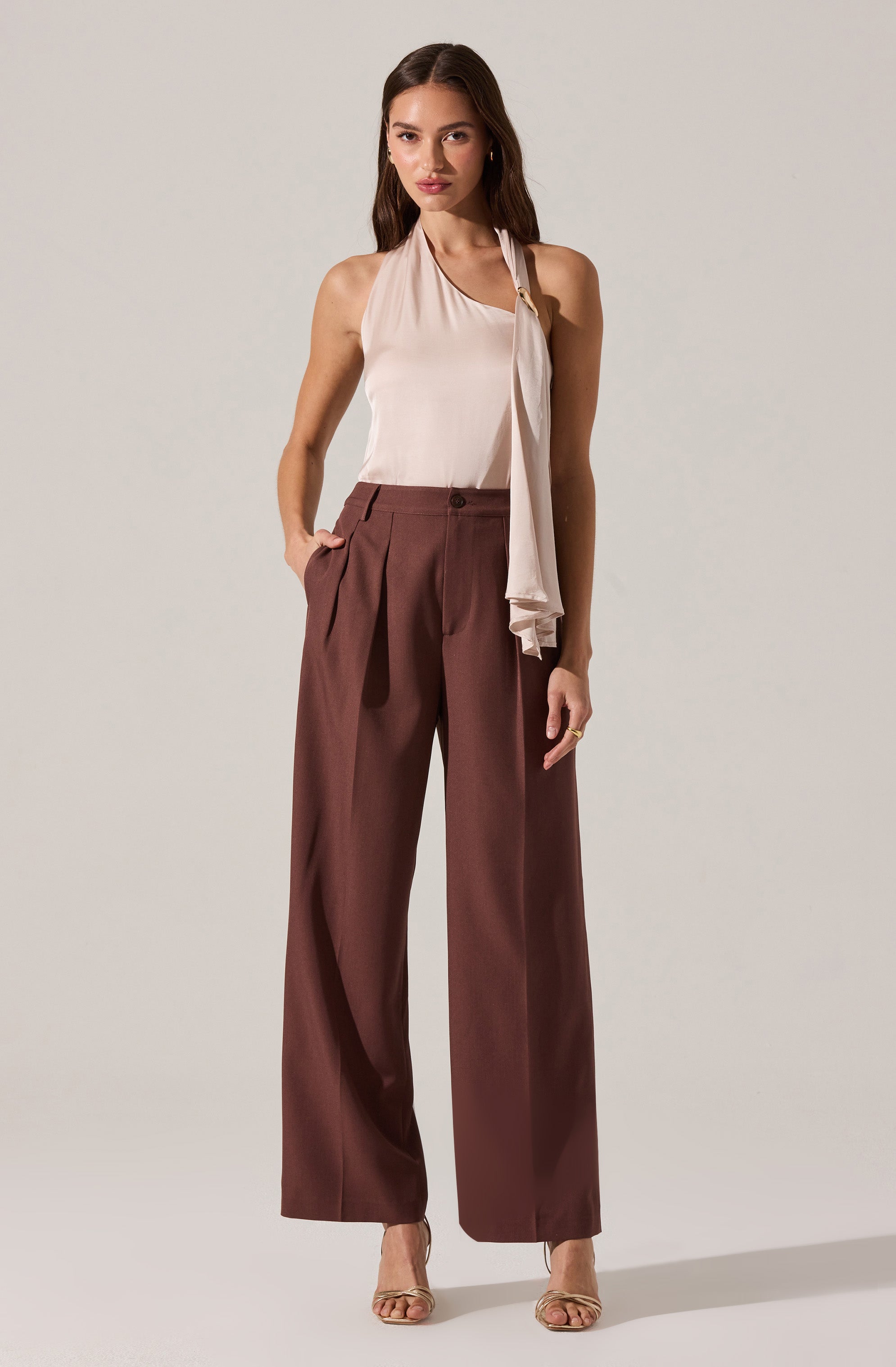 A woman is wearing a sleeveless, light beige blouse with a draped scarf detail hanging from the neckline. She pairs the top with high-waisted, wide-leg brown trousers that feature front pleats and a single button closure. She is also wearing nude, open-toed heels.