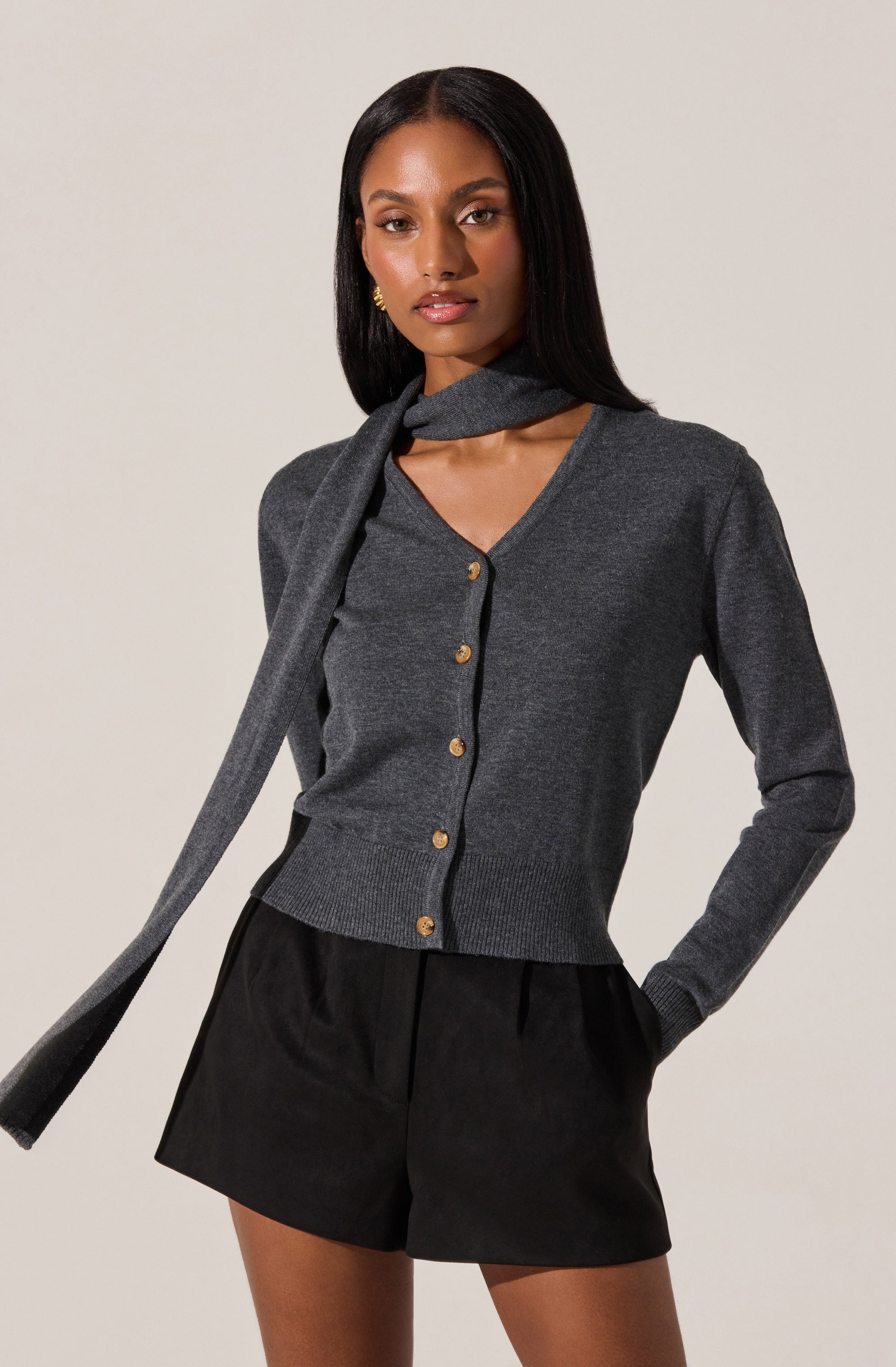 A woman is wearing a dark gray cardigan with gold buttons, paired with a matching gray scarf draped around her neck. She has black shorts and is accessorized with small gold earrings. The outfit presents a sophisticated and elegant look.