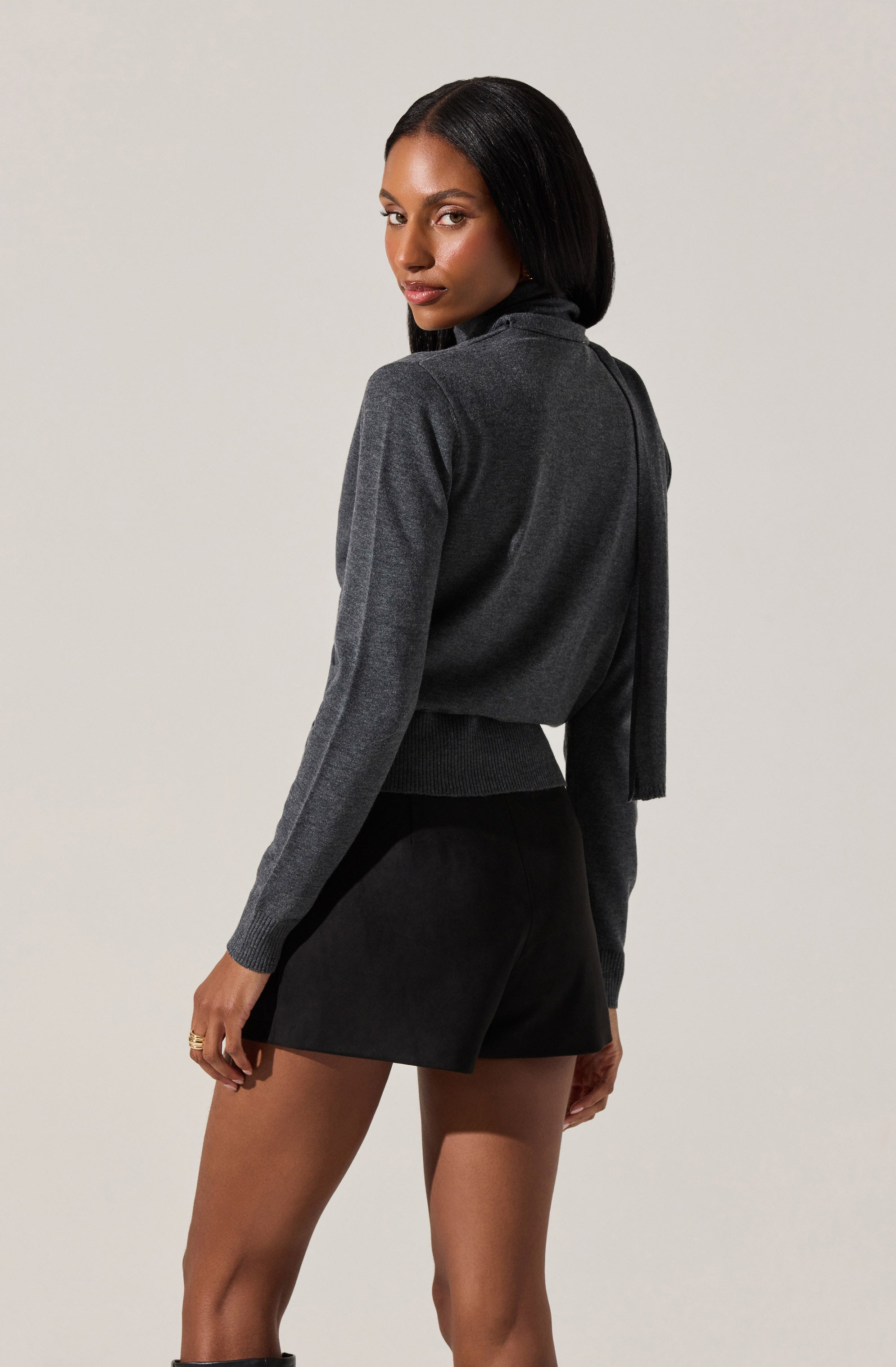 A woman is wearing a dark gray, long-sleeved sweater with a fitted waist and a high neckline. She has paired it with a black skirt that is above the knee and has a sleek, smooth texture.