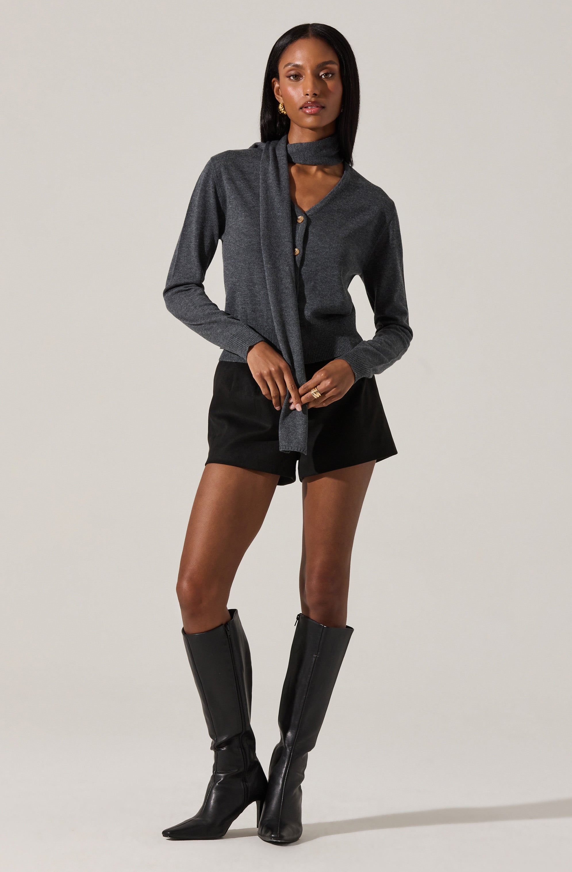 The woman is wearing a dark gray, long-sleeved cardigan with a button-up front. She has a matching dark gray scarf tied around her neck, with the ends hanging down in front. She is also wearing black shorts and knee-high black leather boots with a zipper on the inside. She has gold earrings and a gold ring on her right hand.