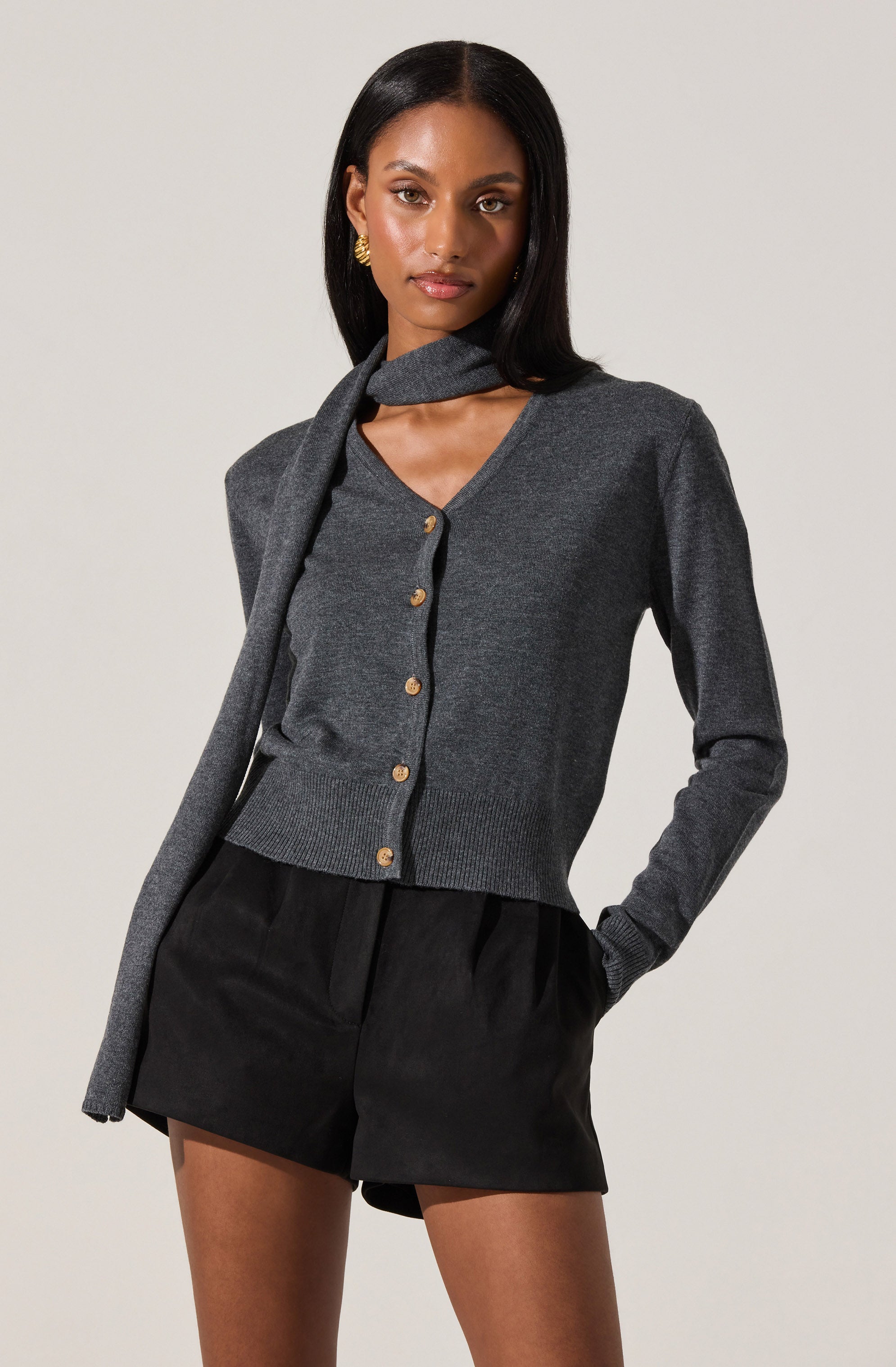 A woman is wearing a dark gray, long-sleeved, button-up cardigan with gold buttons. She has a matching gray scarf draped over her shoulders, with one end hanging in front. She is also wearing high-waisted black shorts with pleats, and has her hands in her pockets. She has dark, straight hair and is accessorized with gold earrings.