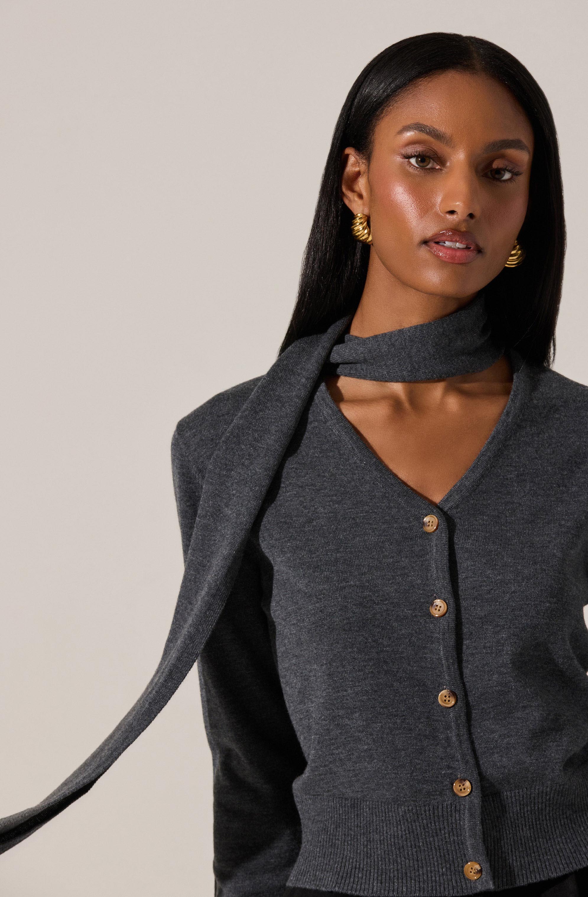A woman is wearing a dark gray cardigan with a V-neckline and wooden buttons down the front. She has a matching dark gray sweater draped over her shoulders as a scarf. She is also wearing gold hoop earrings.