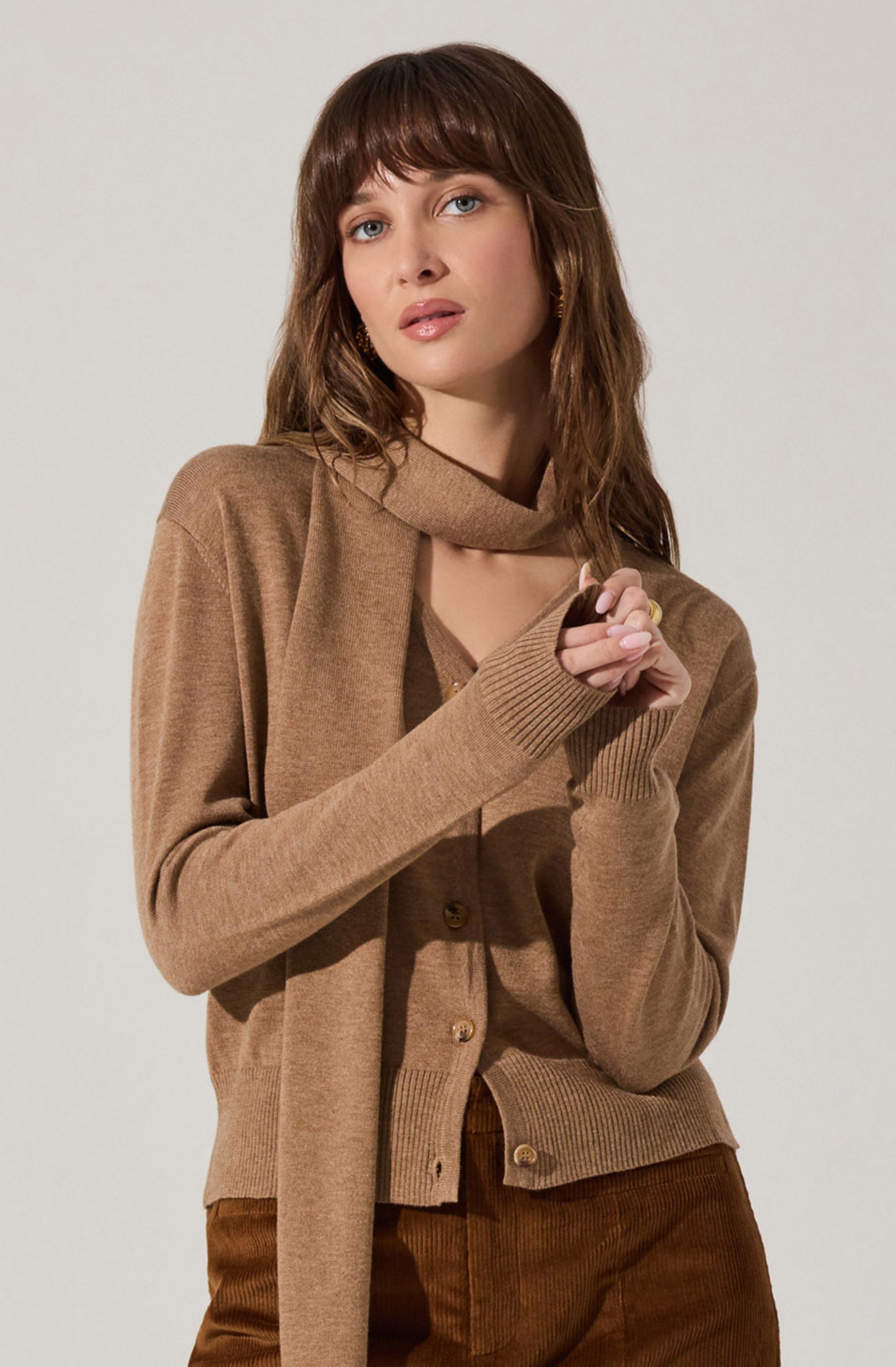 A woman is wearing a beige cardigan sweater with a matching scarf around her neck. Underneath, she has a beige top. She is also wearing brown pants. The overall outfit has a cozy, monochromatic look in shades of beige and brown.