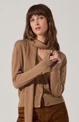 A woman is wearing a beige cardigan sweater with a matching scarf around her neck. Underneath, she has a beige top. She is also wearing brown pants. The overall outfit has a cozy, monochromatic look in shades of beige and brown.