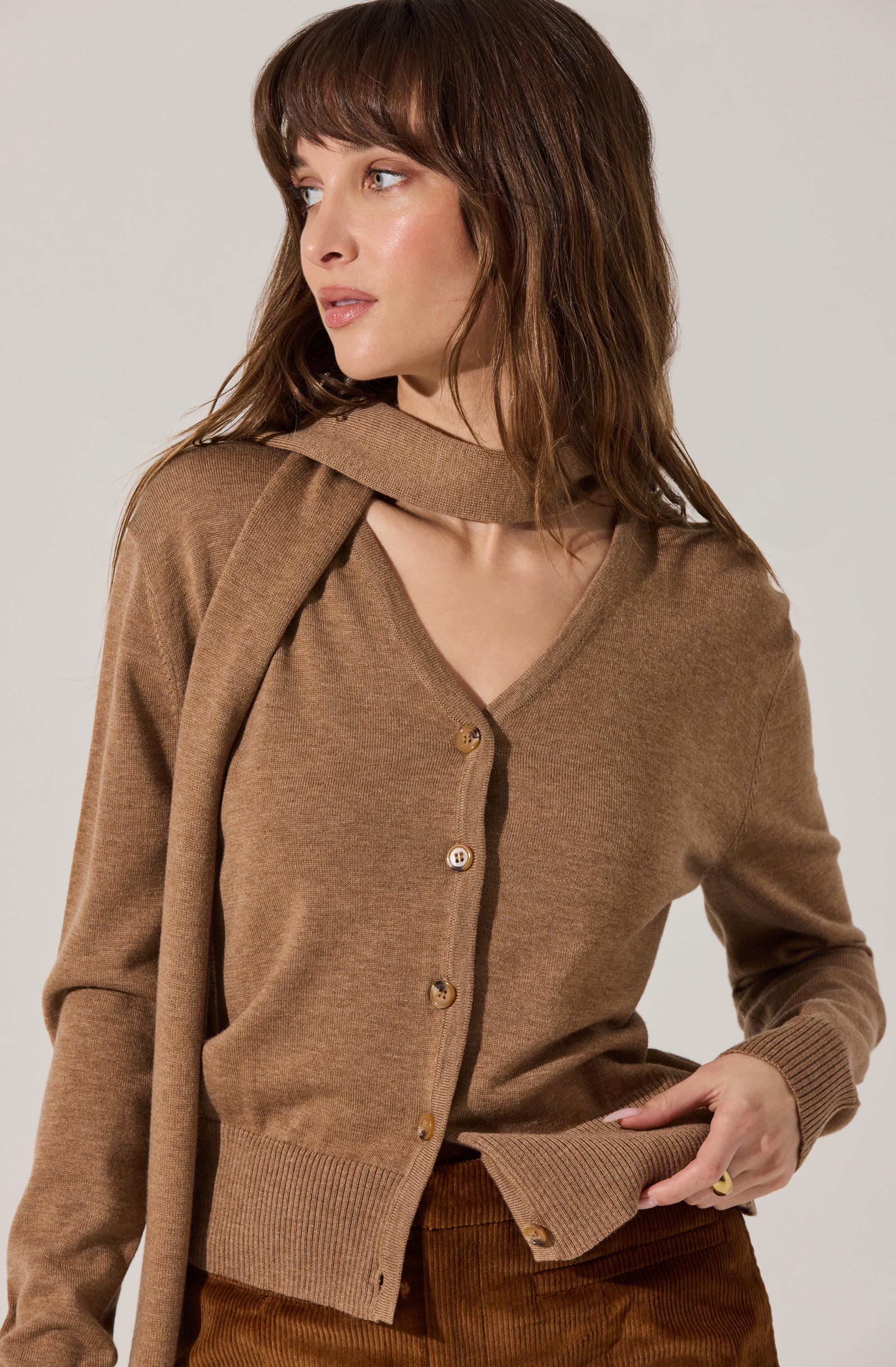 A woman is wearing a camel-colored cardigan with a V-neckline and large buttons down the front. She has a matching scarf draped over her shoulders. Her outfit also includes high-waisted, brown corduroy pants.
