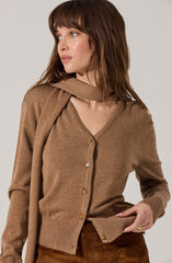 A woman is wearing a camel-colored cardigan with a V-neckline and large buttons down the front. She has a matching scarf draped over her shoulders. Her outfit also includes high-waisted, brown corduroy pants.