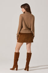 A woman is wearing a light brown cardigan with a high collar and long sleeves, paired with a short brown suede skirt. She has on tall brown suede boots with high heels that reach just below her knees. The outfit is coordinated in various shades of brown.