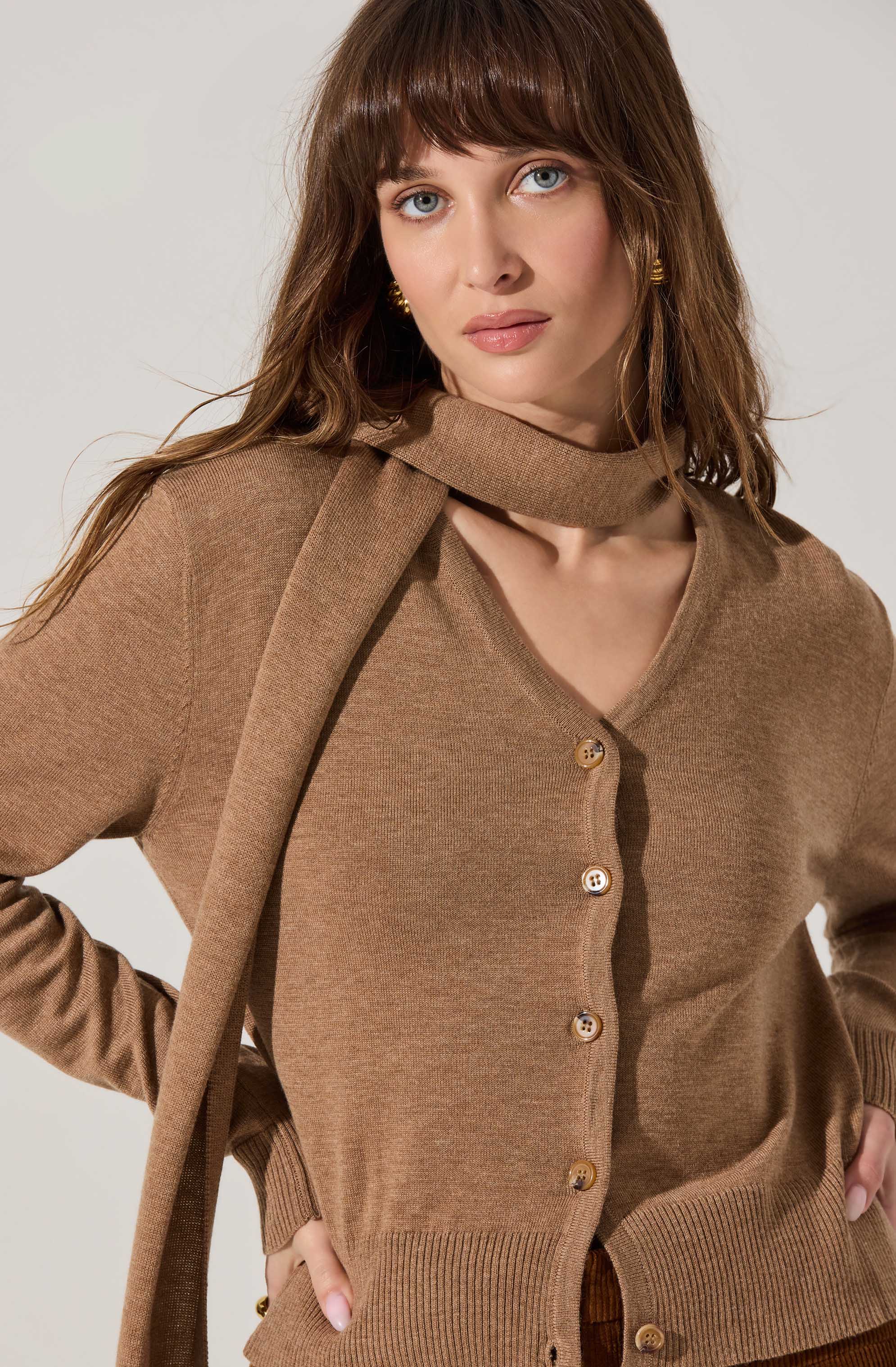 A woman with long brown hair and blue eyes is wearing a brown button-up cardigan with a V-neckline, paired with a matching brown scarf draped around her neck.