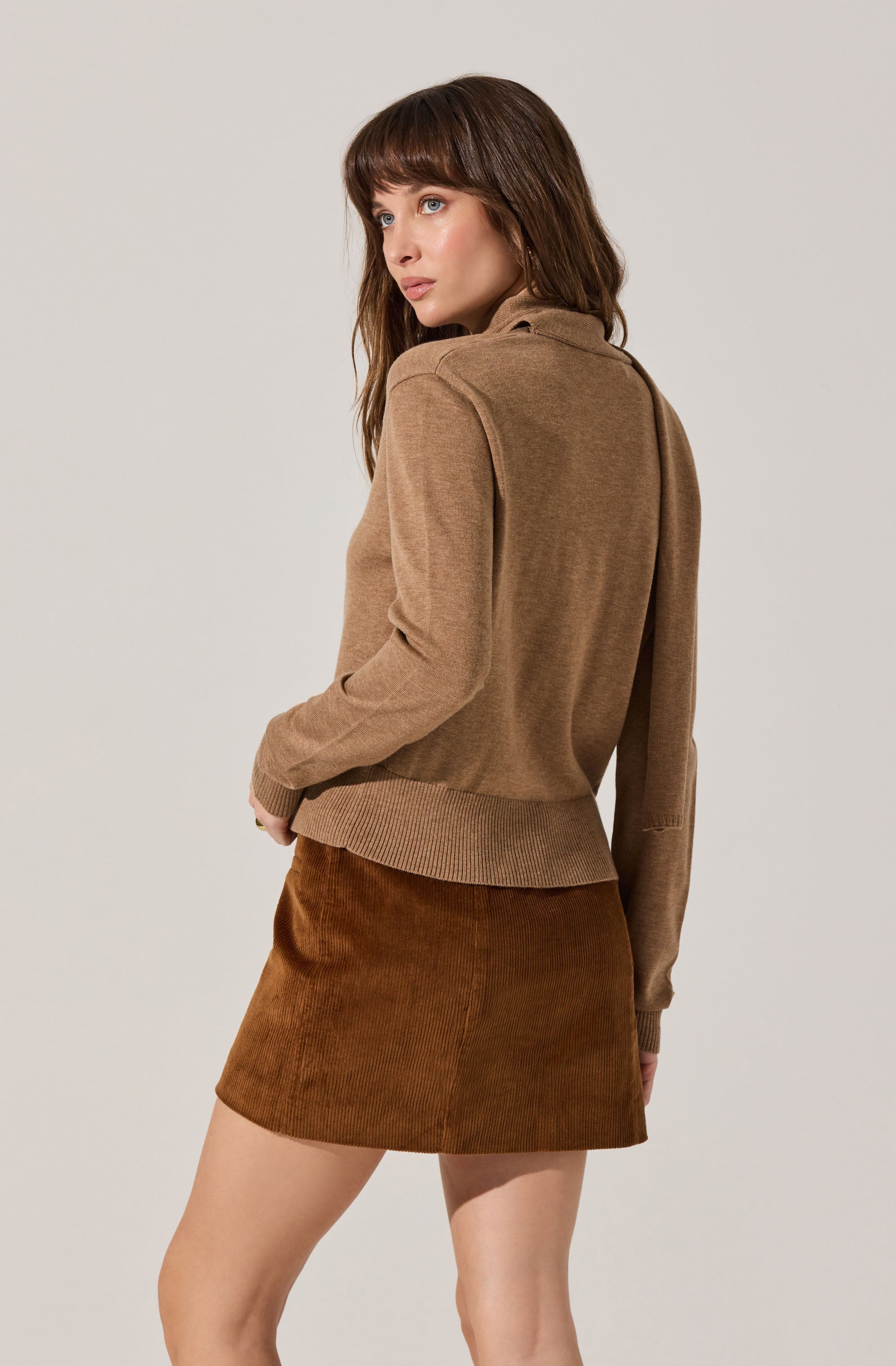 A woman is wearing a light brown, long-sleeved sweater with a high collar and ribbed hem, paired with a brown corduroy skirt. The sweater has a relaxed fit, and the skirt is a mini length.