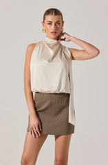 A woman is wearing a sleeveless, high-neck, satin-like cream-colored top with draping details. She pairs it with a high-waisted, fitted mini skirt featuring a small checkered pattern in shades of brown. She accessorizes with gold hoop earrings and a statement ring.