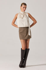 A woman is wearing a sleeveless, cream-colored satin blouse with a high neckline and a large, draped bow hanging from the collar. She pairs it with a high-waisted, brown checkered mini skirt and tall, black knee-high boots. She has her hair styled in a sleek, pulled-back look and is accessorized with gold earrings.