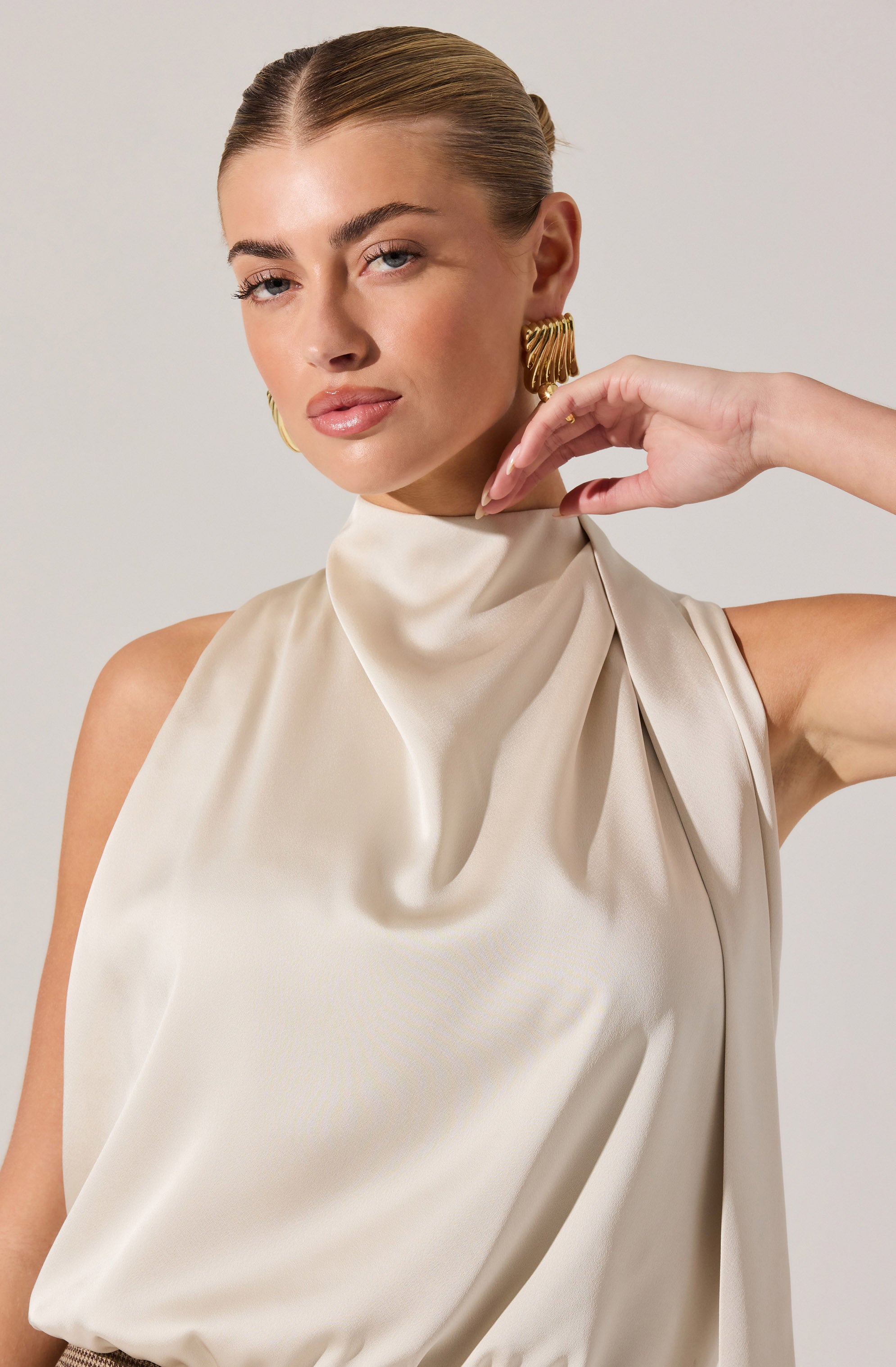 A woman wears a sleeveless, high-neck satin top in an off-white or champagne color. The top has a draped, gathered design at the neckline, creating a soft, elegant look. She is accessorized with large, gold, rectangular hoop earrings.