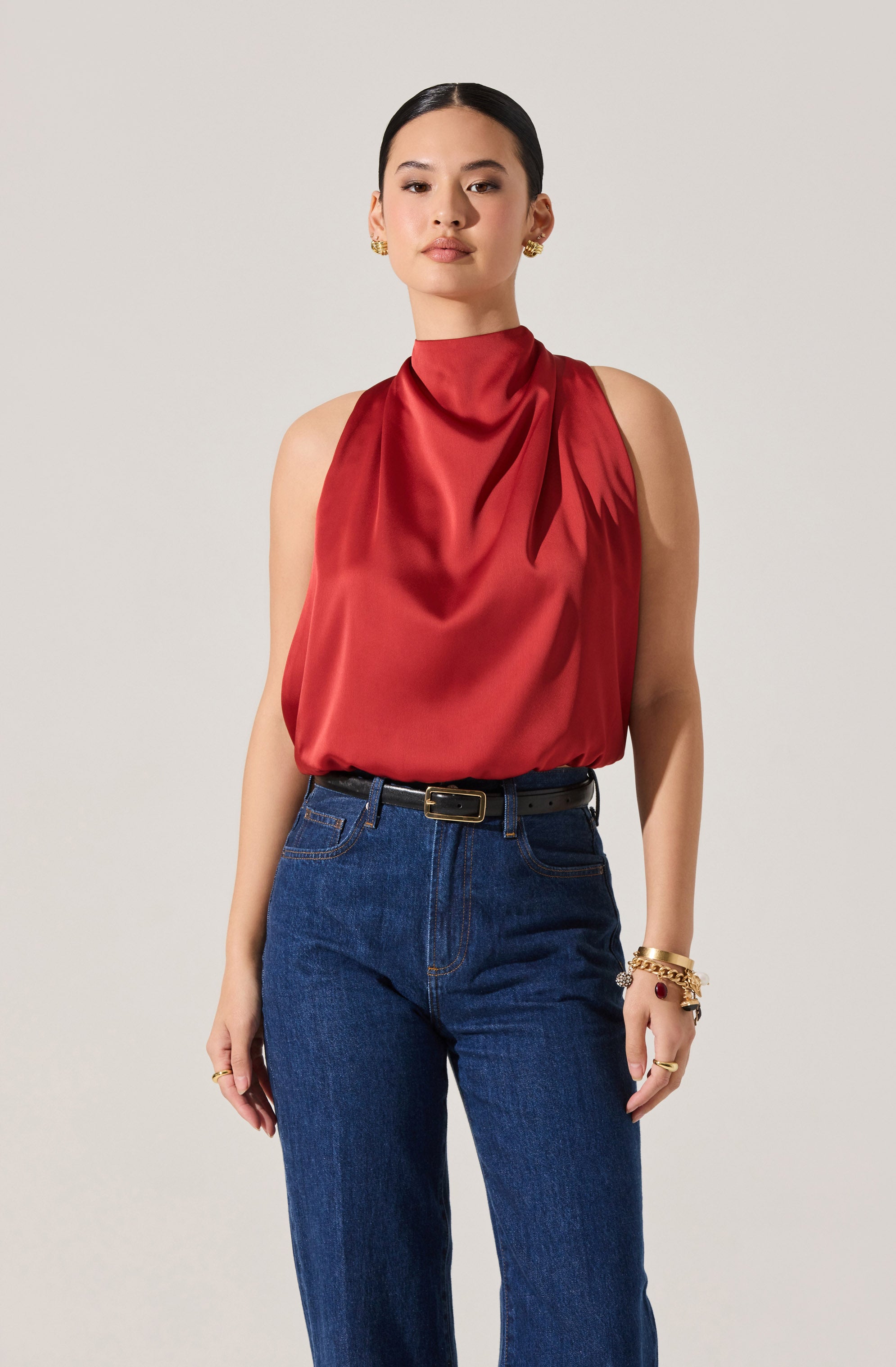 A woman is wearing a sleeveless, high-neck satin top in a rich red color with soft draping at the front. She pairs it with high-waisted, dark blue denim jeans.