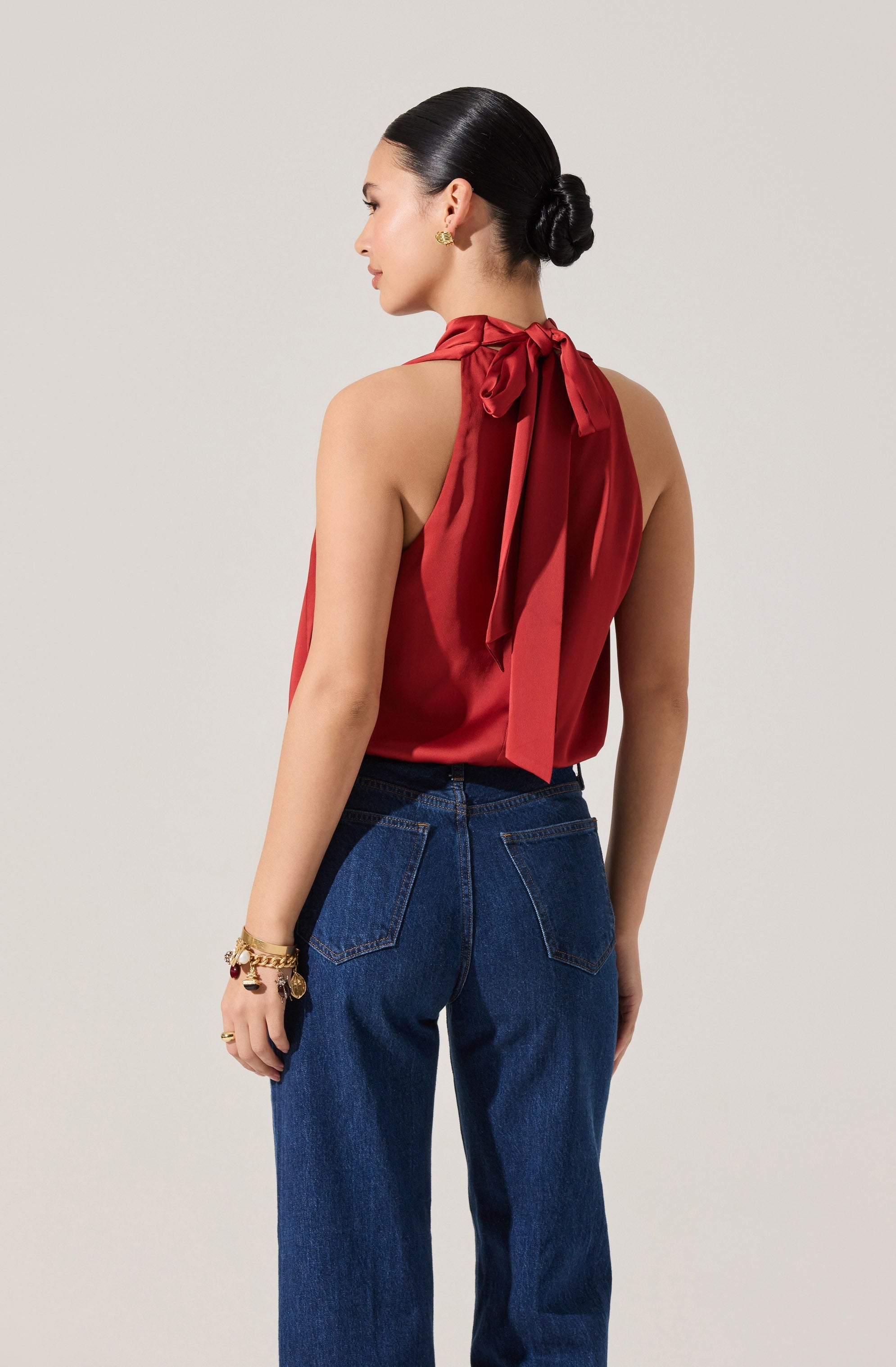 A woman is wearing a sleeveless, red satin top with a high neckline that features a large bow tied at the back. She pairs it with high-waisted, dark blue denim jeans. She accessorizes with gold jewelry, including earrings and bracelets.
