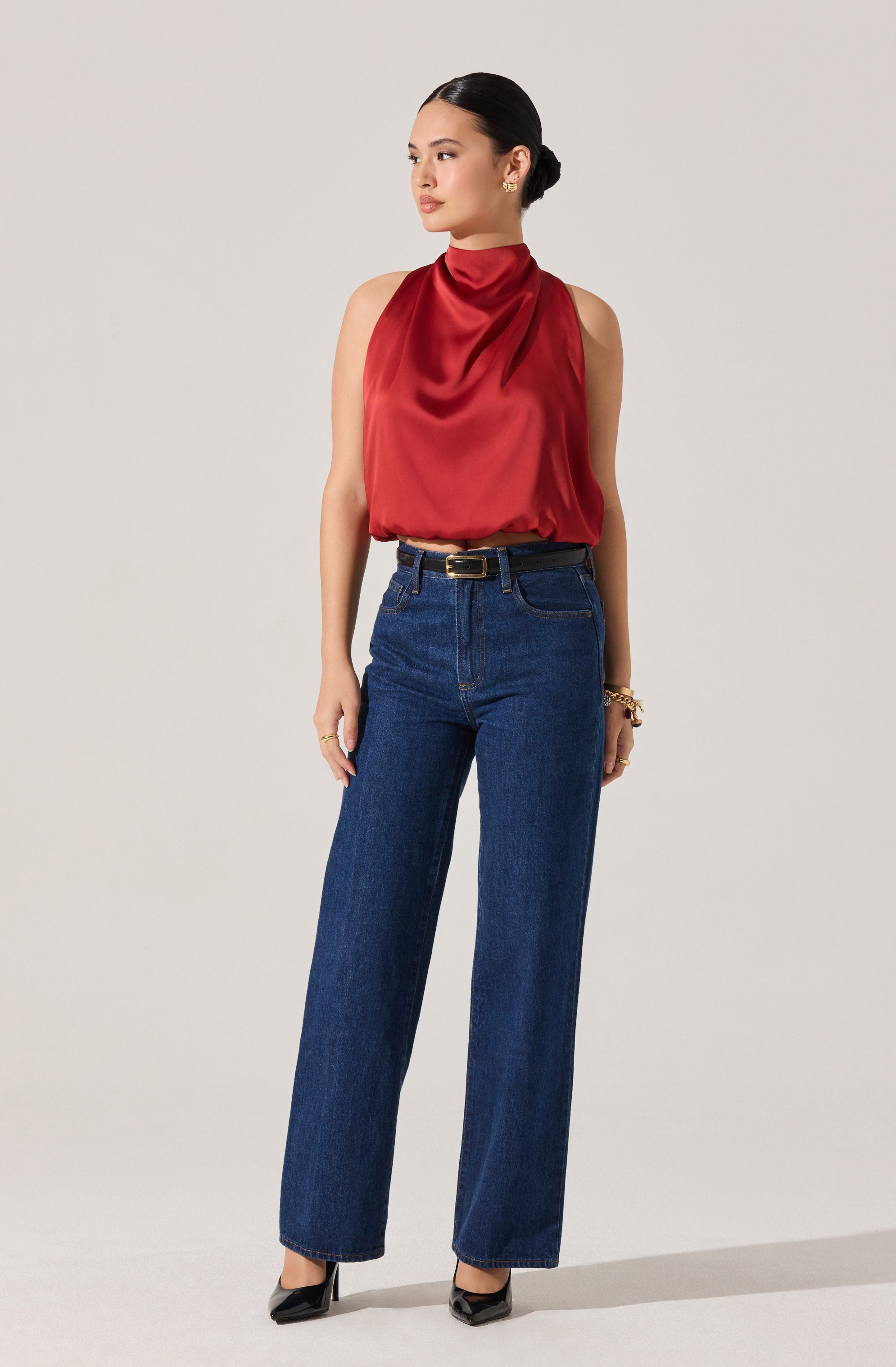 A woman is wearing a sleeveless, high-neck, silky red top that drapes softly around her neck and torso. She has paired it with high-waisted, straight-leg blue jeans and a black belt with a gold buckle. She is also wearing black pointed-toe heels.