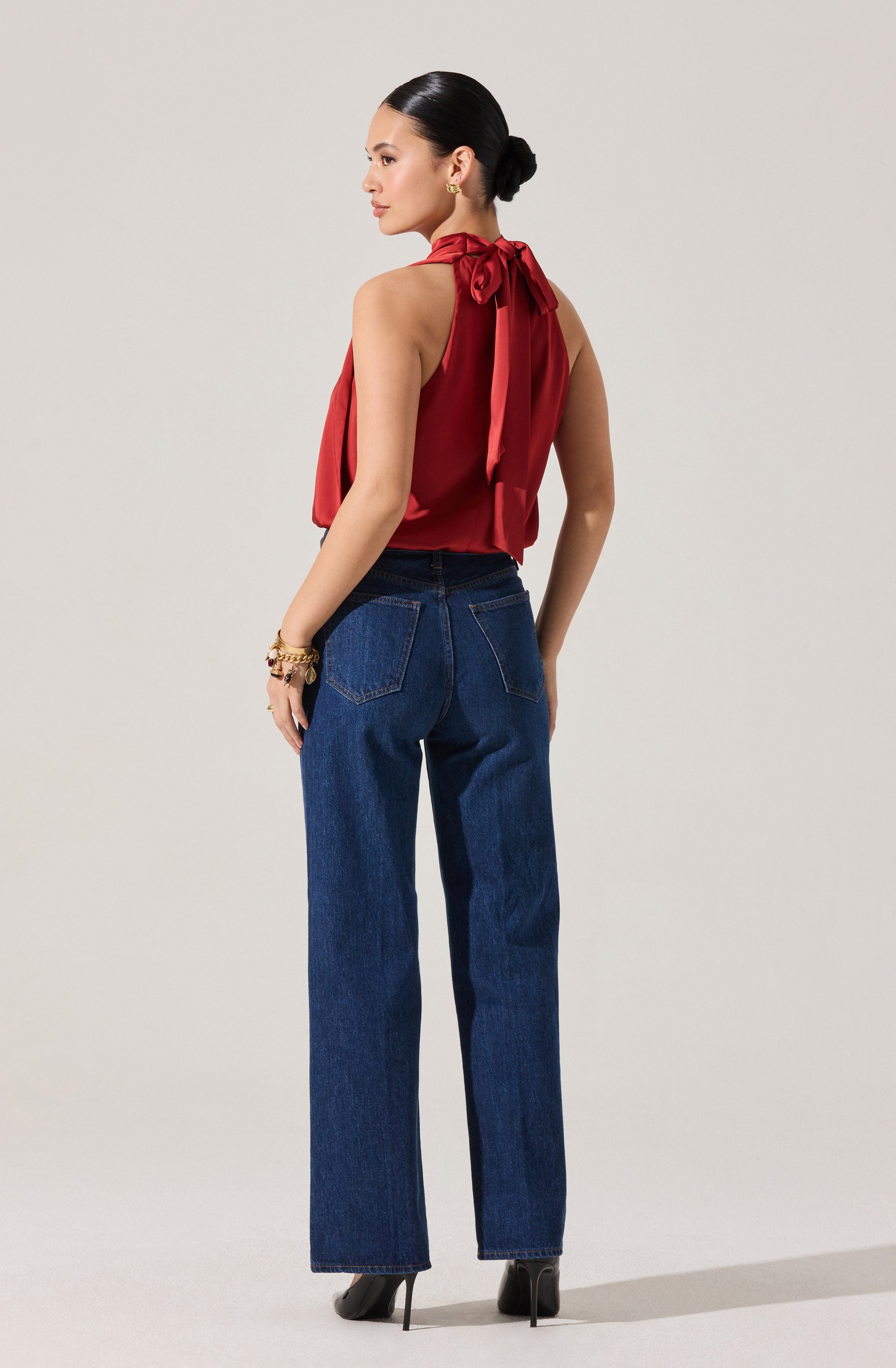 A woman is wearing a sleeveless, red top with a high neckline that ties into a bow at the neck. She is also wearing high-waisted, straight-leg blue jeans and black high-heeled shoes. She has her hair styled in a low bun and is accessorized with multiple bracelets on her right wrist.