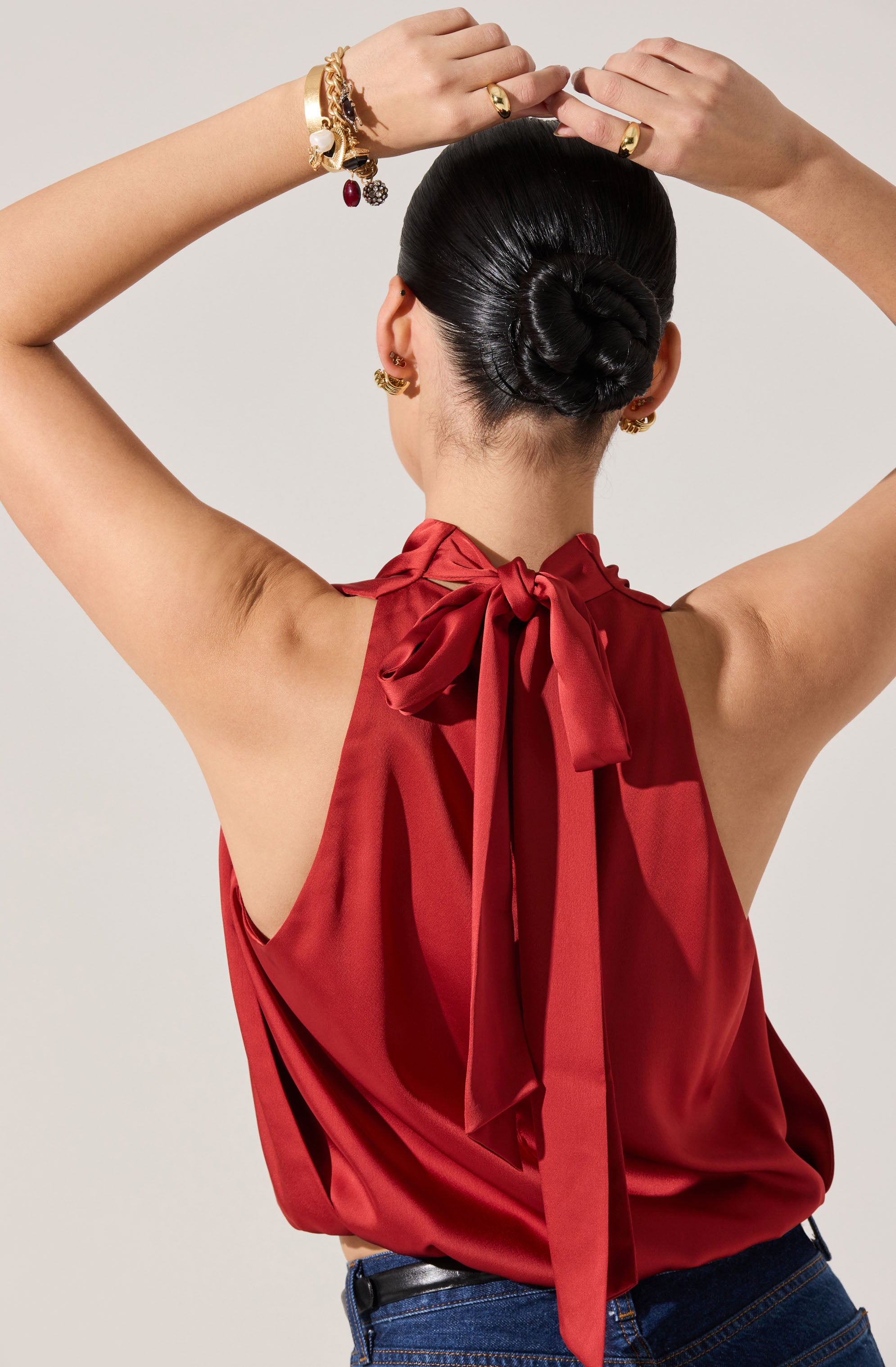 A woman is wearing a sleeveless red satin top with a large bow tied at the back of the neckline. She is also wearing dark blue jeans. Her hair is styled in a sleek, low bun, and she has multiple gold earrings and bracelets, some adorned with charms and stones.