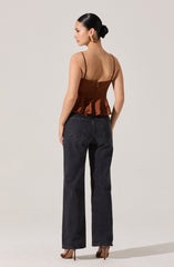 A woman is wearing a sleeveless brown satin top with thin spaghetti straps and a ruffled hem, paired with high-waisted, straight-leg black jeans. She has her hair styled in a low bun and is accessorizing with large gold earrings.