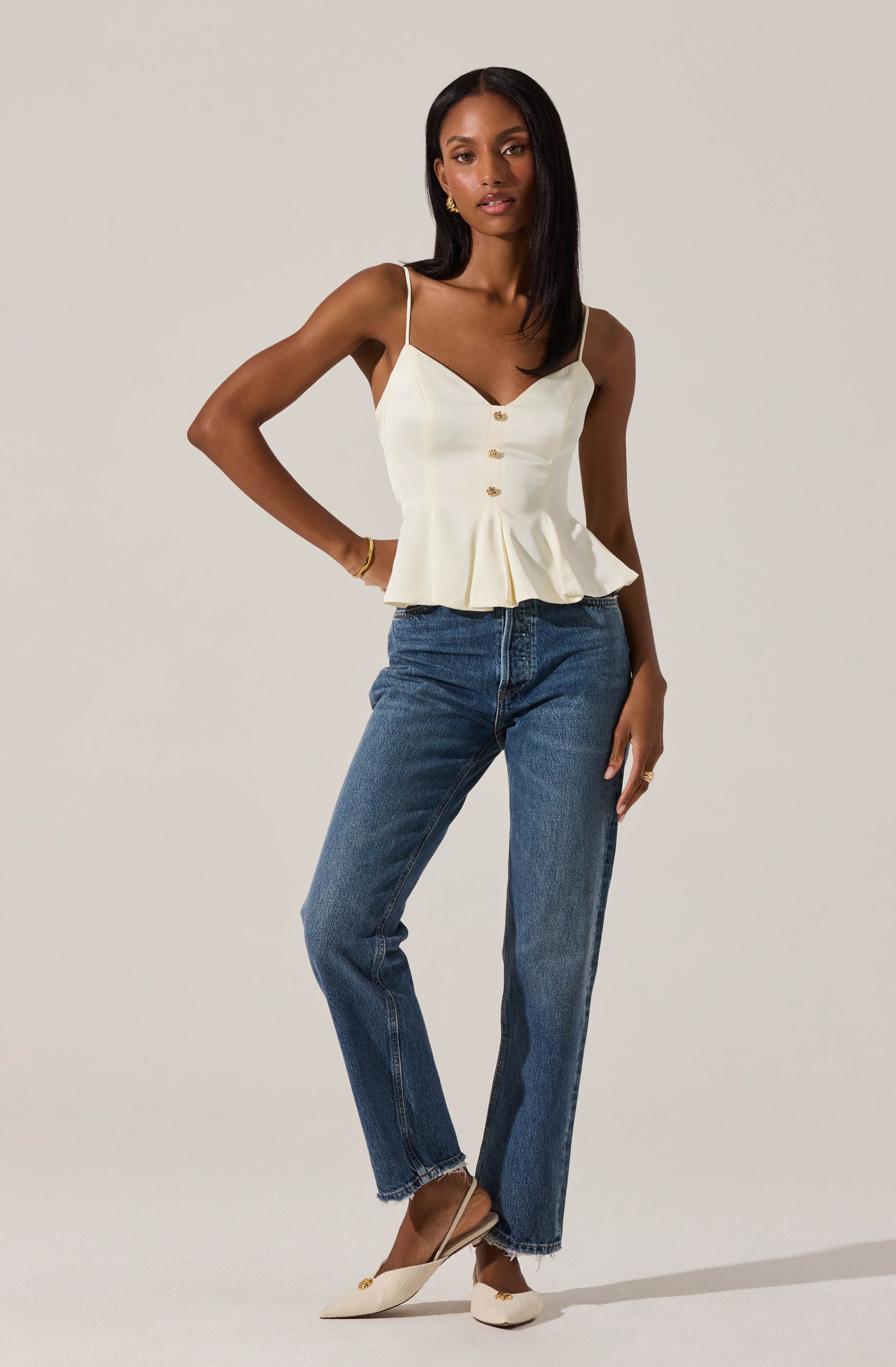 A woman is wearing a white sleeveless top with thin spaghetti straps, featuring a V-neckline and button details down the front, along with a flared hem. She pairs it with high-waisted, straight-leg blue jeans and beige pointed-toe heels.