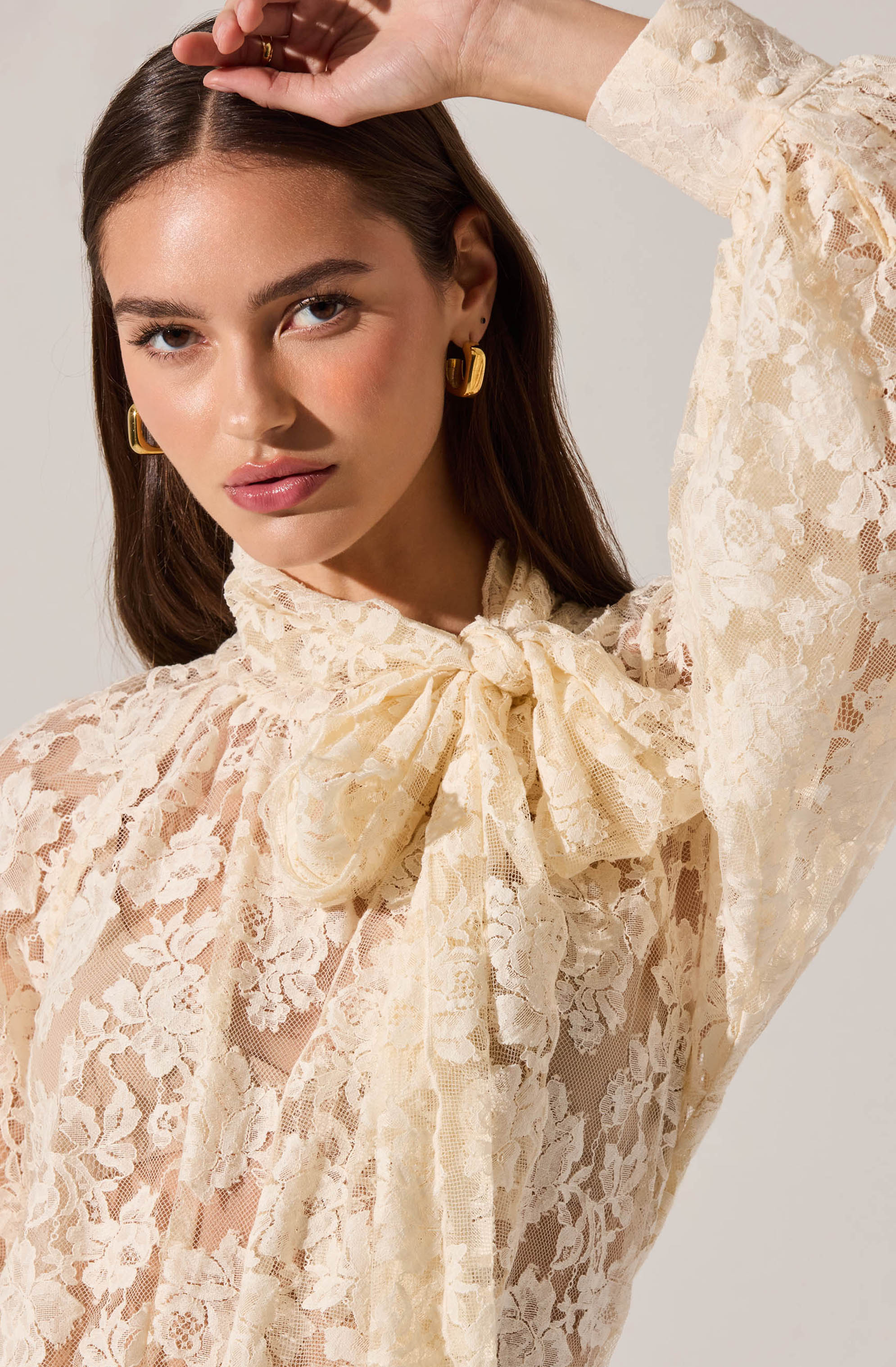 A woman is wearing a cream-colored lace blouse with a high neck and a large bow at the collar. The blouse features intricate floral lace patterns and sheer fabric. She is accessorized with gold hoop earrings.
