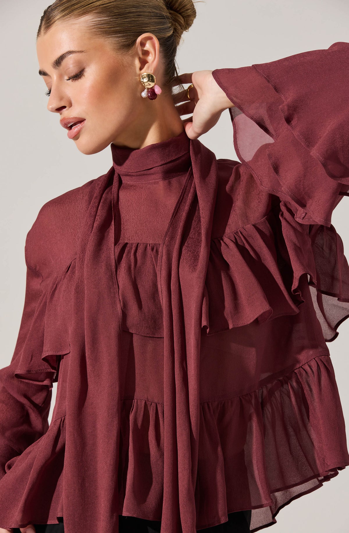 A woman is wearing a dark red, semi-sheer blouse with a high neckline. The blouse features ruffled details around the shoulders and chest, with ruffles extending down the front and along the sleeves. The fabric appears lightweight and flowy, creating a soft and feminine look. She is accessorizing with large, ornate earrings with gold, pearl, and gemstone elements.