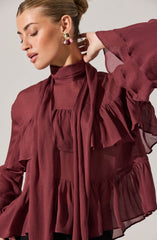 A woman is wearing a dark red, semi-sheer blouse with a high neckline. The blouse features ruffled details around the shoulders and chest, with ruffles extending down the front and along the sleeves. The fabric appears lightweight and flowy, creating a soft and feminine look. She is accessorizing with large, ornate earrings with gold, pearl, and gemstone elements.