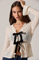 A woman wearing a cream-colored, long-sleeved, lace blouse with a textured, open-knit pattern. The blouse features a ruffled neckline and is adorned with two black velvet bows in the front, creating a decorative tie. She is also wearing dark blue jeans. Her hair is long and brown, and she has on gold earrings.