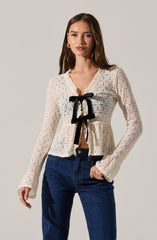 The woman is wearing a long-sleeved, cream-colored, open-knit cardigan with a scalloped edge along the neckline. The cardigan features a decorative black bow tied at the front center, just below the bust. She is also wearing dark blue jeans.