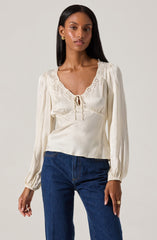 A woman is wearing a long-sleeved, cream-colored satin blouse with a V-neckline trimmed with eyelet lace and a small tie detail at the center. The blouse features puffed shoulders and gathered cuffs. She pairs it with dark blue jeans.