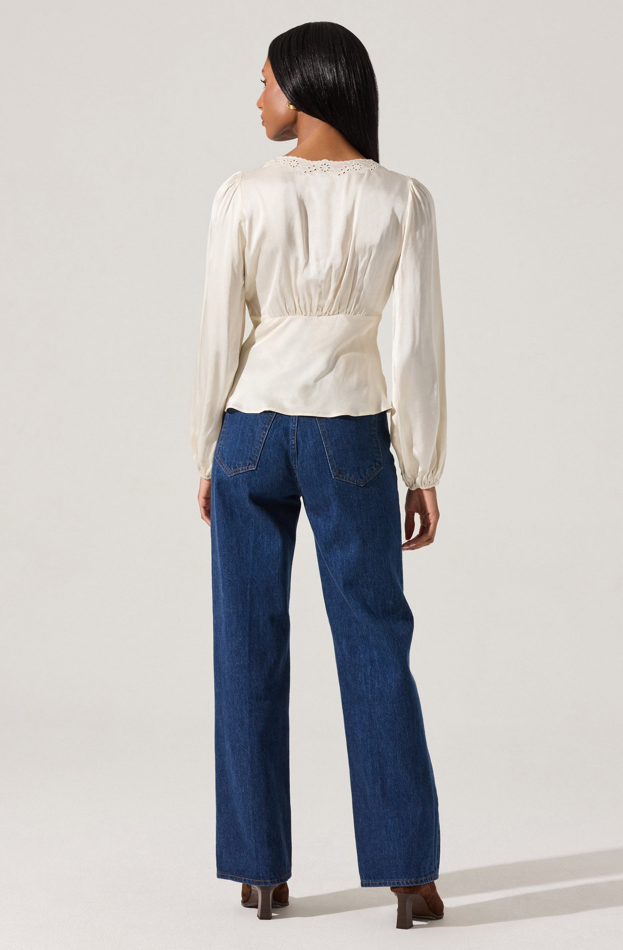A woman is wearing a long-sleeved, cream-colored blouse with a subtle sheen and gathered waist, featuring delicate eyelet embroidery along the neckline. She is paired with high-waisted, straight-leg blue jeans and brown heeled shoes.