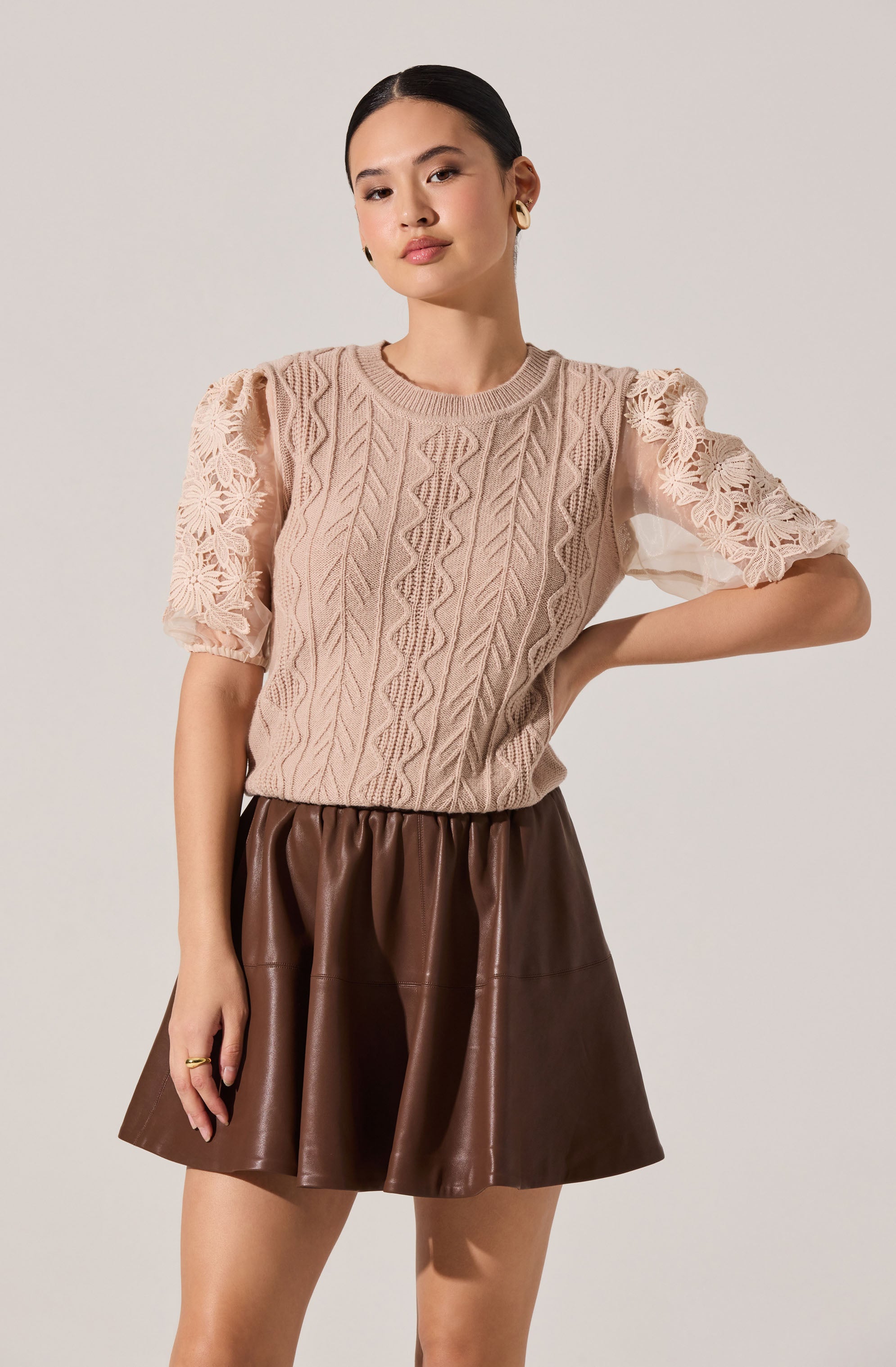 A woman is wearing a beige, knit sweater with a cable and leaf pattern, featuring semi-sheer, puffed lace sleeves with floral detailing. She pairs it with a high-waisted, dark brown, faux leather skirt that is slightly flared.
