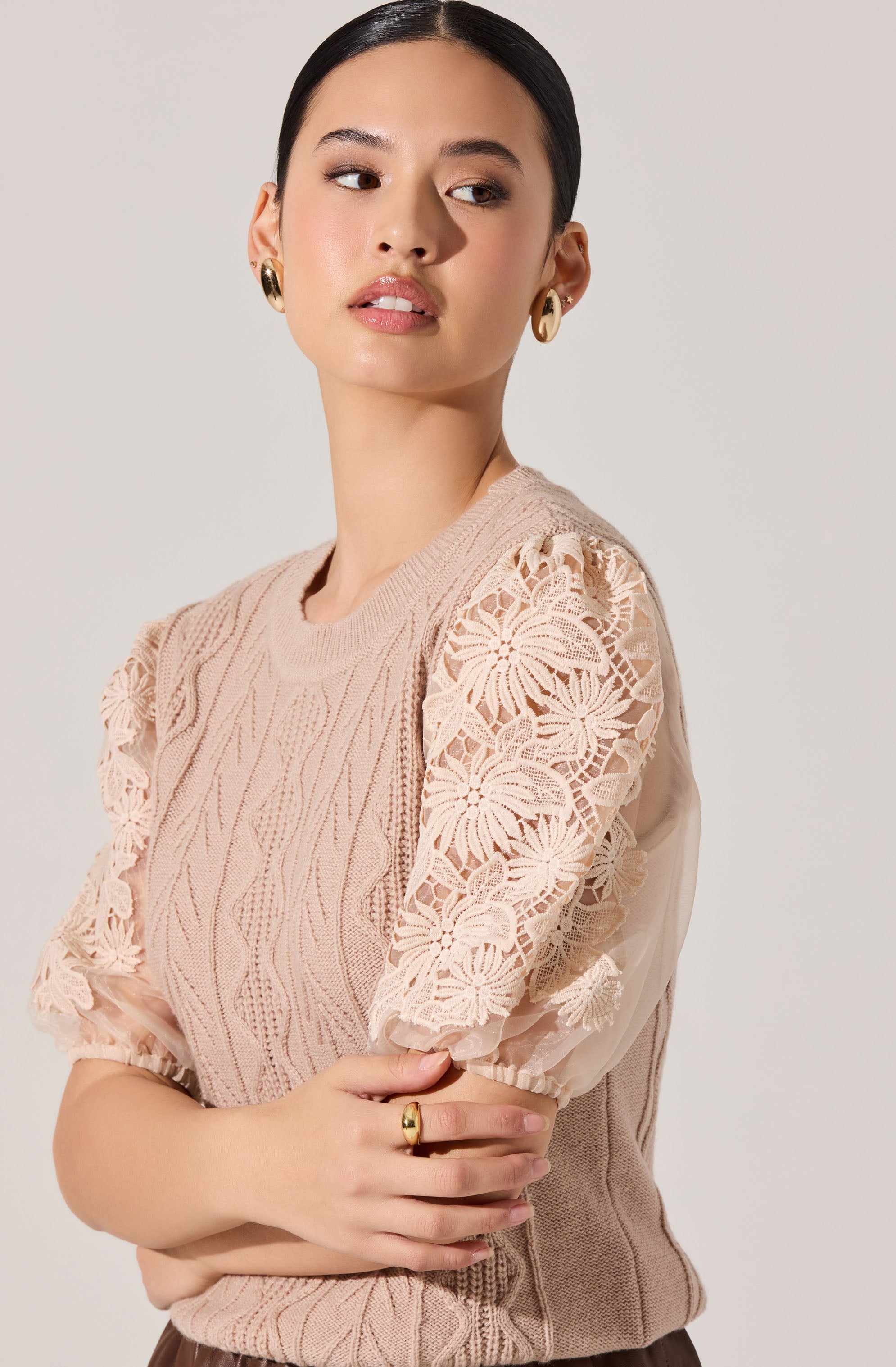 A woman with dark hair pulled back is wearing a beige knitted top with intricate lace detailing on the sleeves, which feature floral and leaf patterns. The top has a textured cable-knit design on the main body. She accessorizes with large gold hoop earrings and a ring on her right hand.