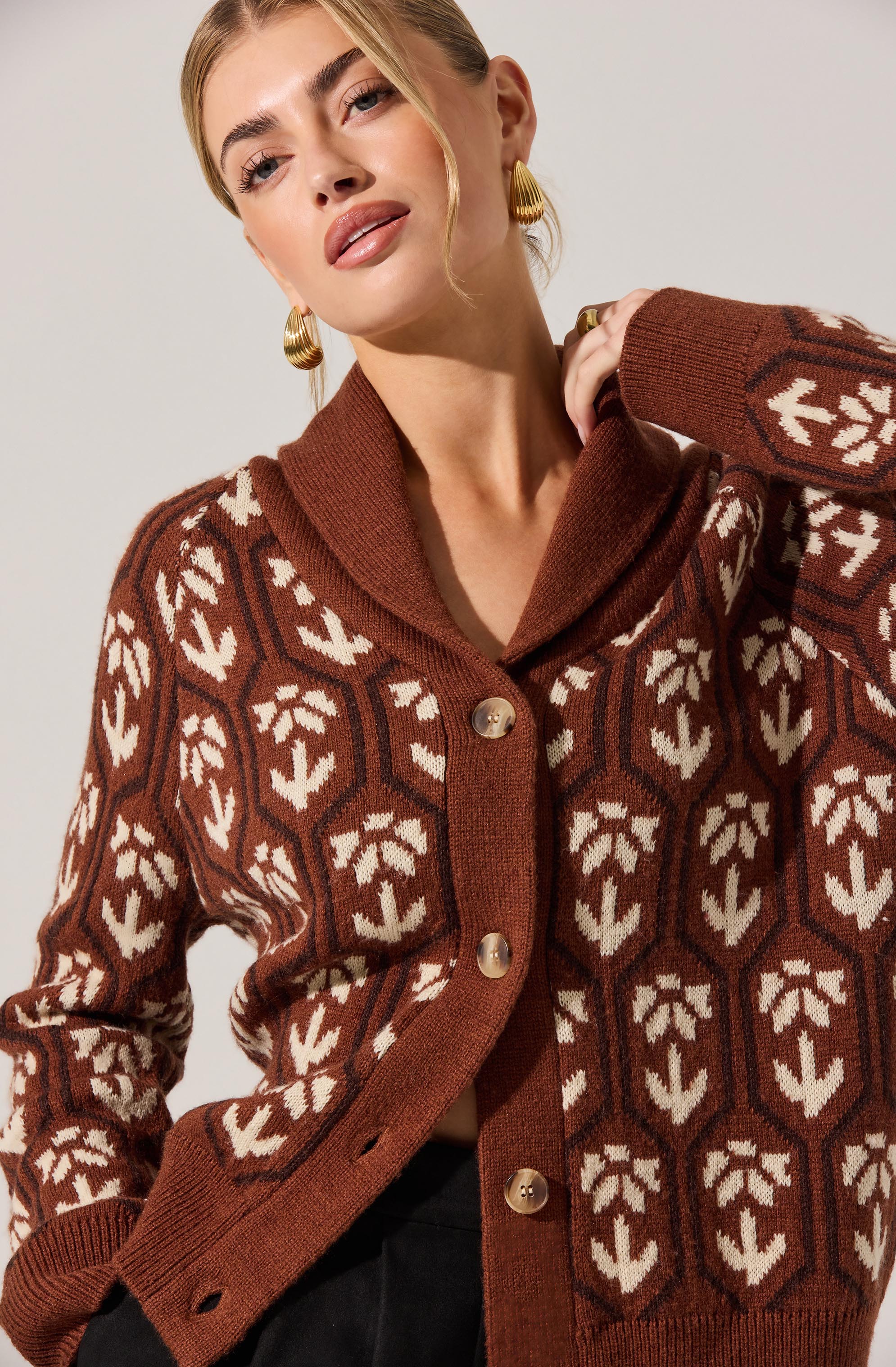 A woman wearing a brown, patterned cardigan with cream-colored leaf and geometric designs. The cardigan features a shawl collar and large buttons down the front.