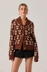 A woman is wearing a brown cardigan with a white geometric pattern throughout. The cardigan has a wide, draped collar and button closures down the front. She is also wearing black shorts with a relaxed fit.
