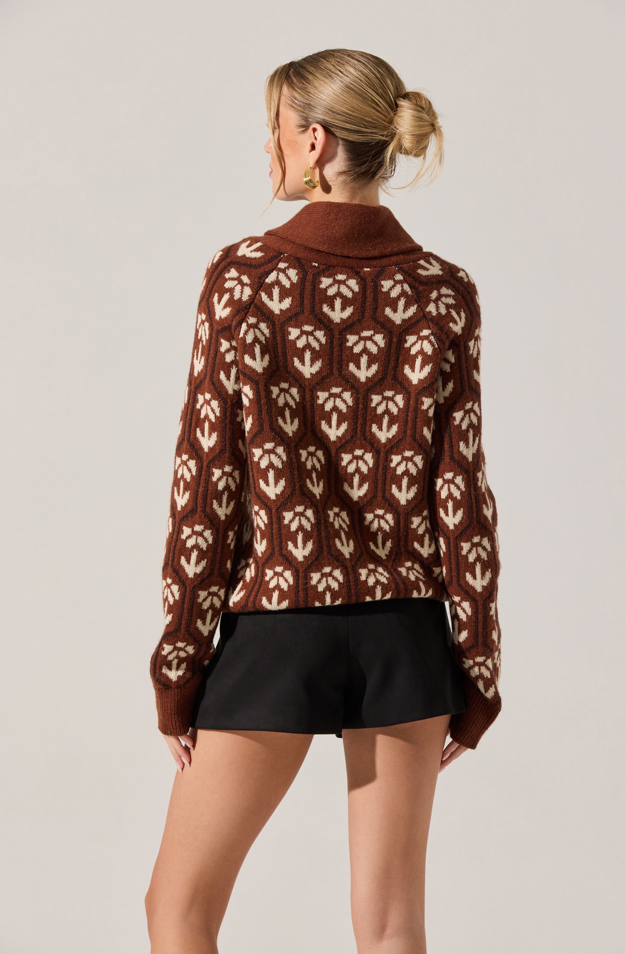 A woman is wearing a brown sweater with a geometric pattern of cream-colored shapes resembling stylized flowers or leaves. The sweater has a wide collar and long sleeves that extend past her wrists. She pairs it with black shorts. Her hair is styled in a low, messy bun.