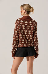 A woman is wearing a brown sweater with a geometric pattern of cream-colored shapes resembling stylized flowers or leaves. The sweater has a wide collar and long sleeves that extend past her wrists. She pairs it with black shorts. Her hair is styled in a low, messy bun.