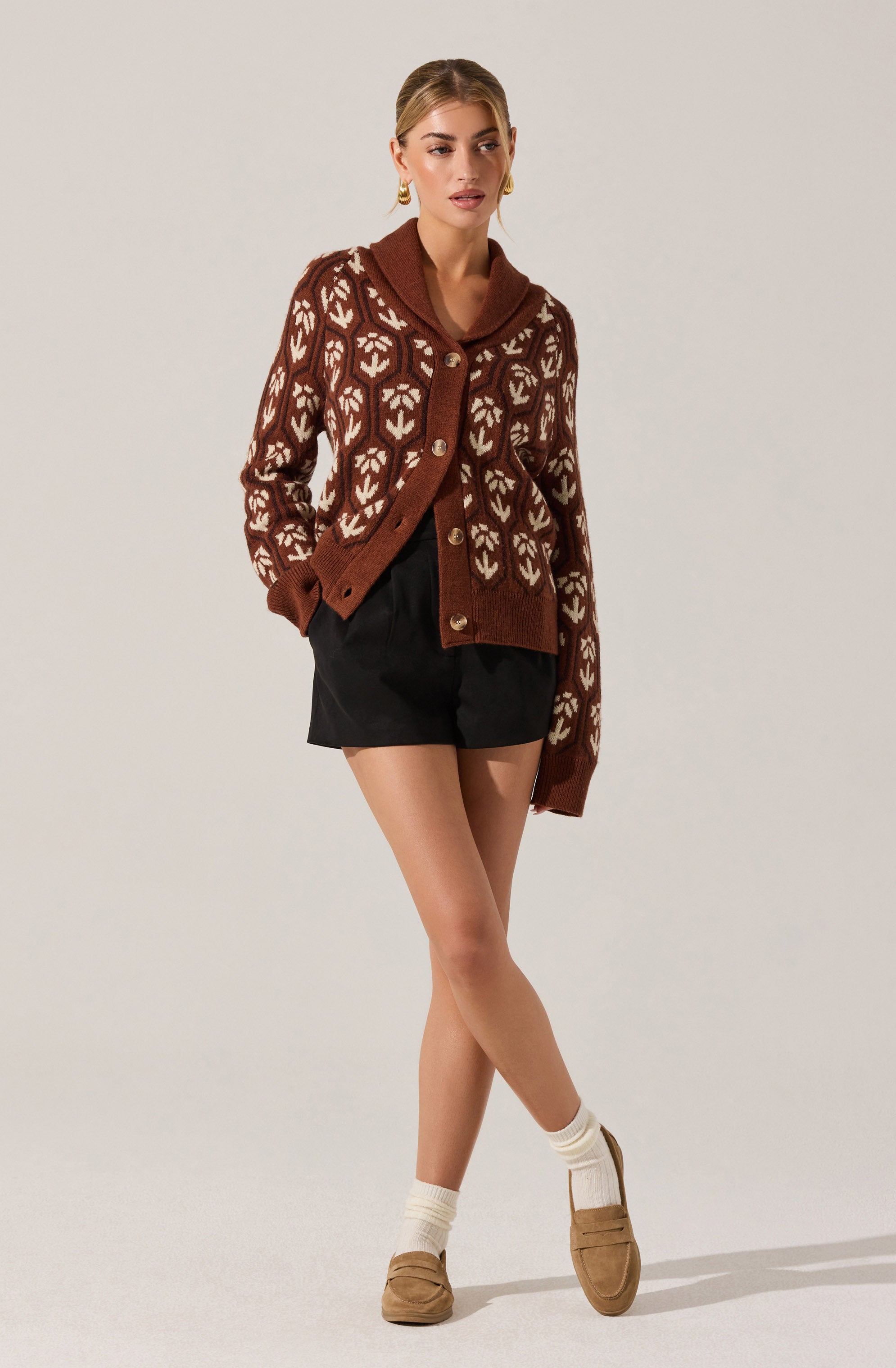 A woman is wearing a brown cardigan with a white leaf-like pattern, large buttons down the front, and a wide collar. She has paired it with black shorts, white socks, and tan loafers.