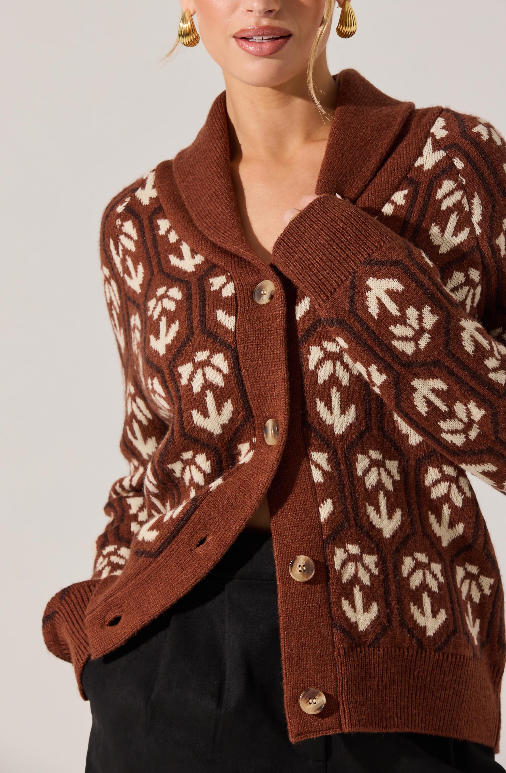 A woman is wearing a brown knitted cardigan with a white geometric leaf pattern throughout. The cardigan has a wide, rounded collar and large, round brown buttons down the front. She has her hand in a pocket, and the sleeve cuffs are ribbed. The cardigan appears to be made of a warm, cozy material.