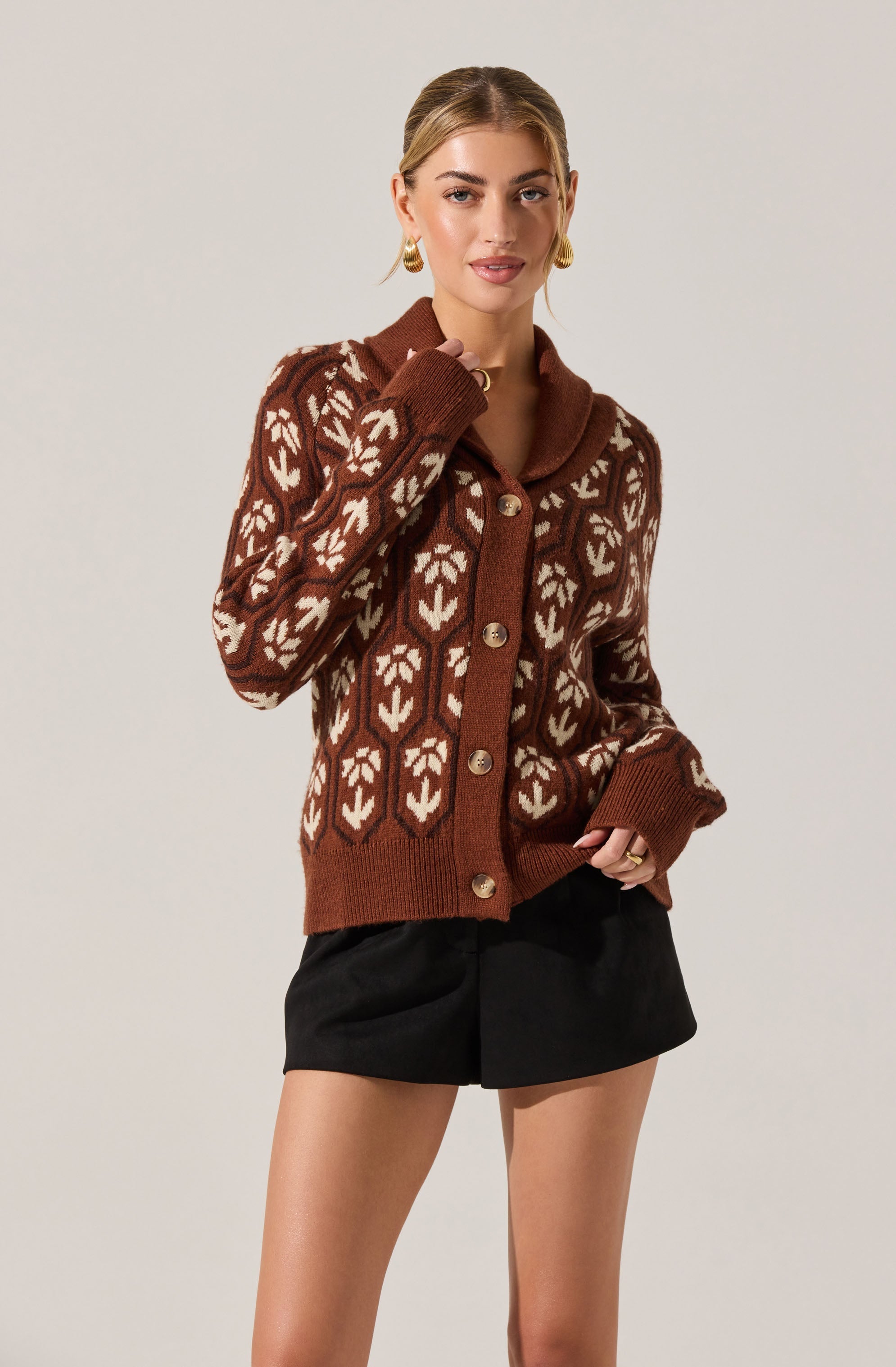 A woman is wearing a brown cardigan sweater with a white leaf or floral pattern. The sweater features large buttons down the front, a wide collar, and long sleeves with ribbed cuffs. She pairs it with black shorts.