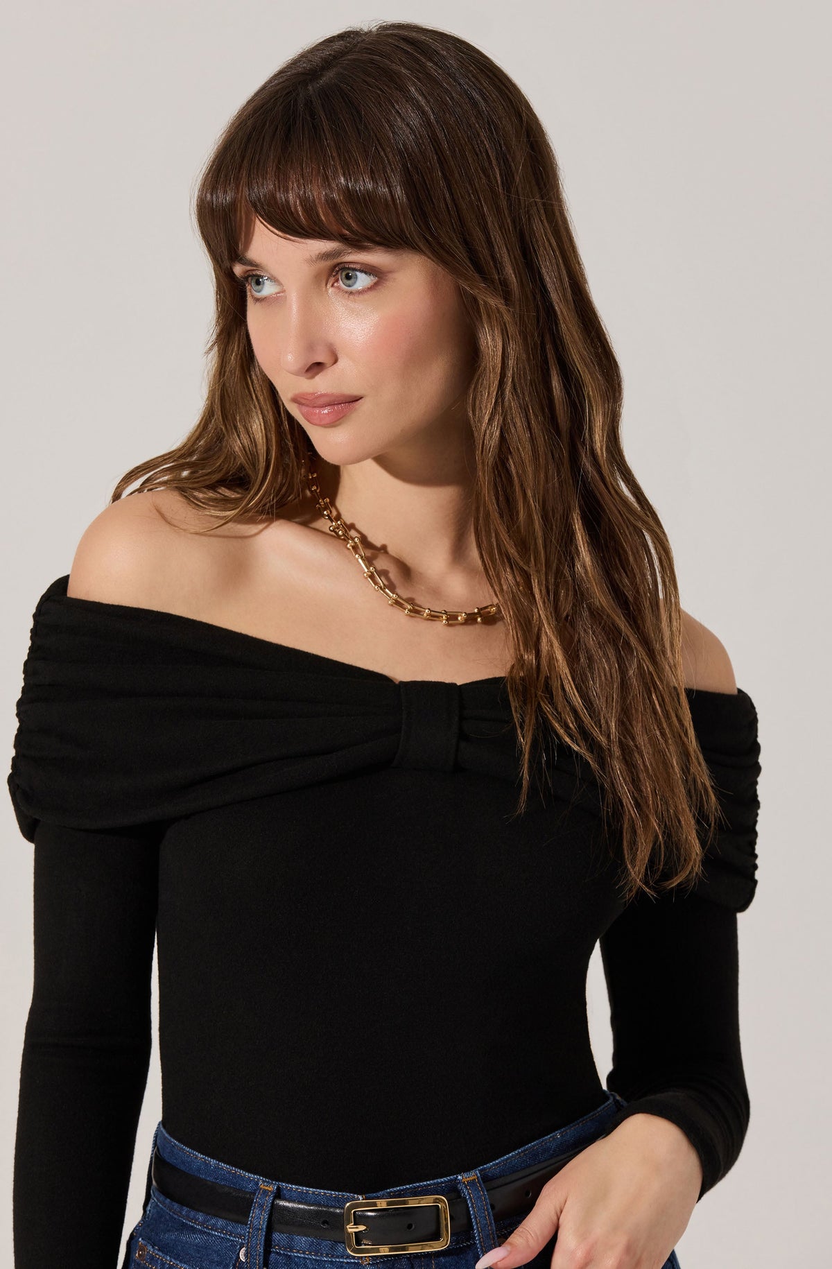 A woman is wearing a black off-the-shoulder top with padded, ruched sleeves and a large bow detail at the neckline. She has paired the top with dark blue jeans and a black belt with a gold buckle. She is also wearing a gold chain necklace.